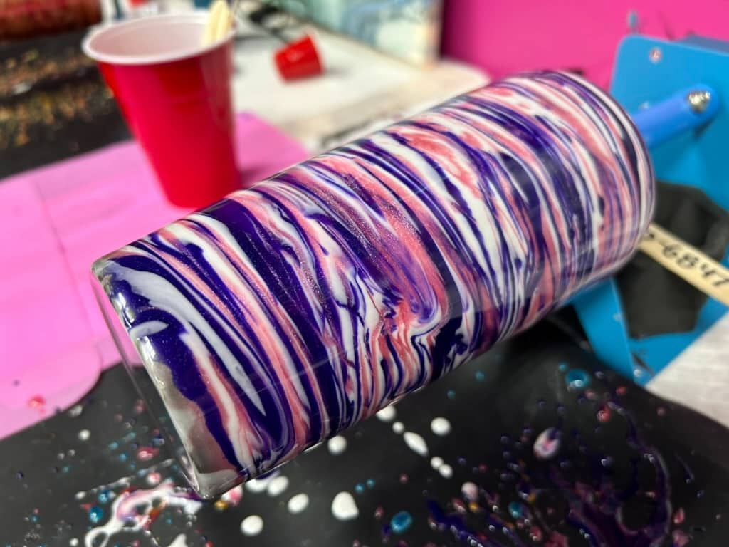 Paint-swirled tumbler on a blue stand, with pink, purple, and white drips on a craft table