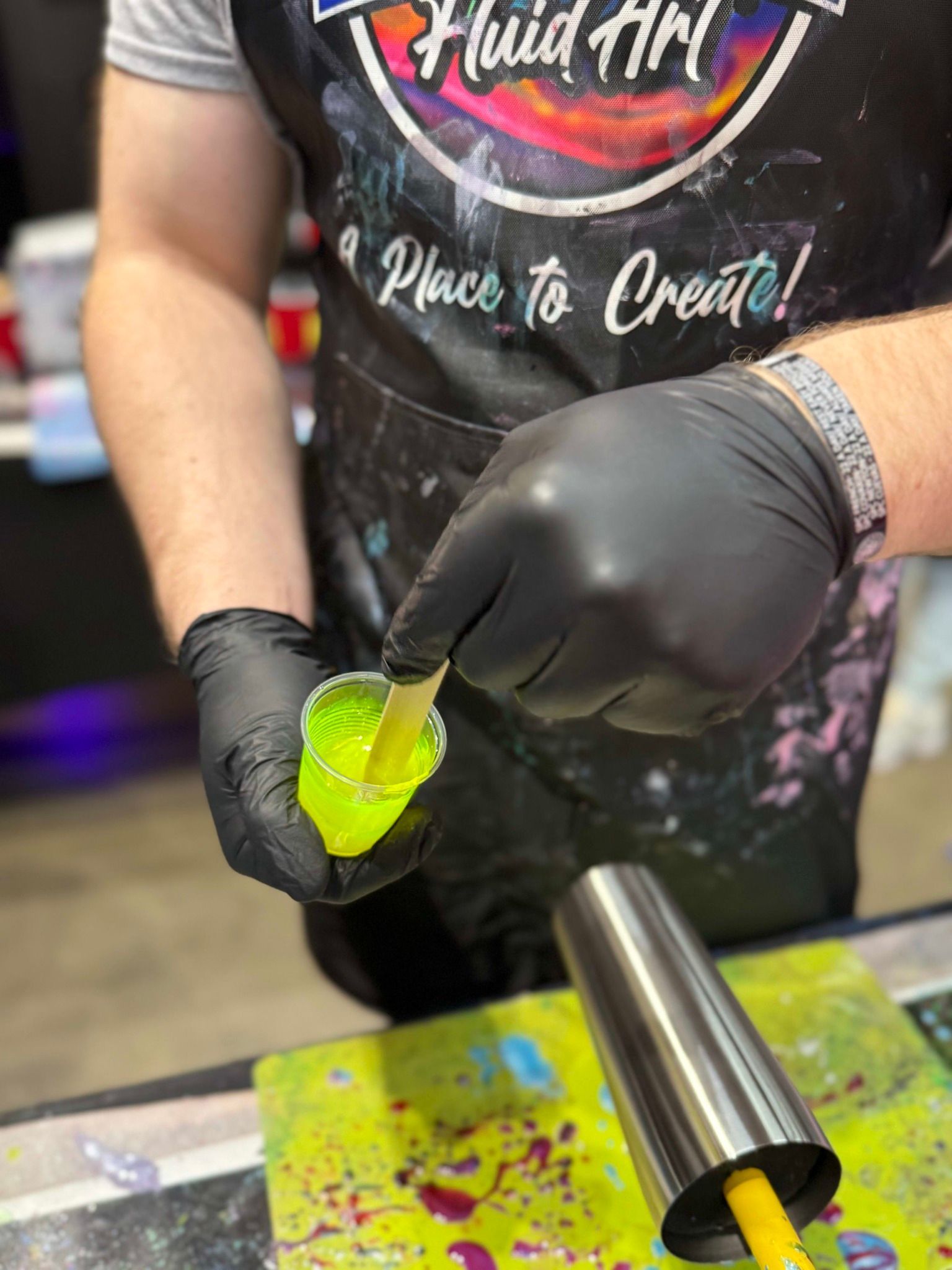 Person in black gloves pours bright green resin into a clear cup at a craft/workshop table.