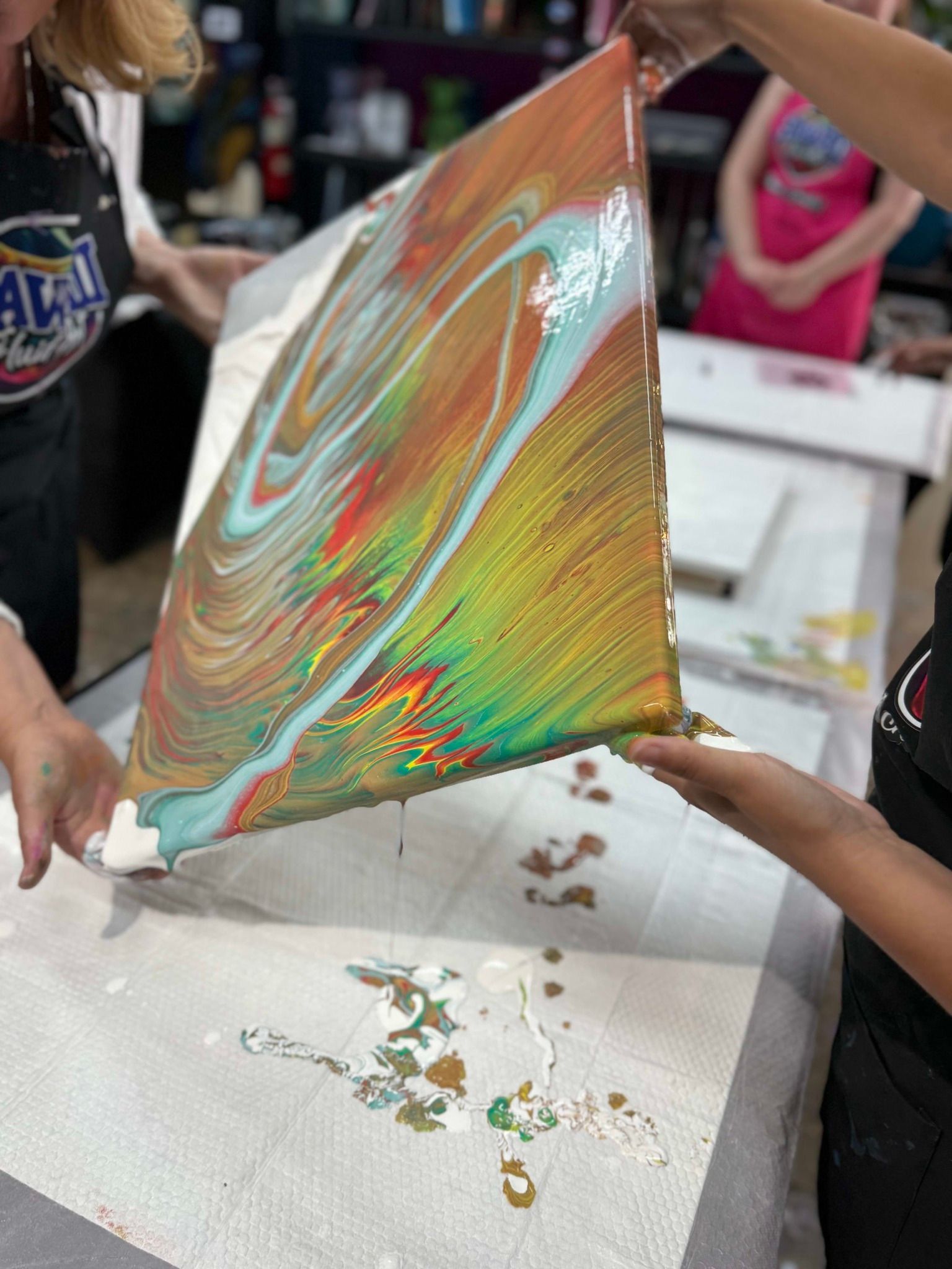 Person lifts a rainbow iridescent sheet over a table with spilled craft liquid in a busy indoor setting