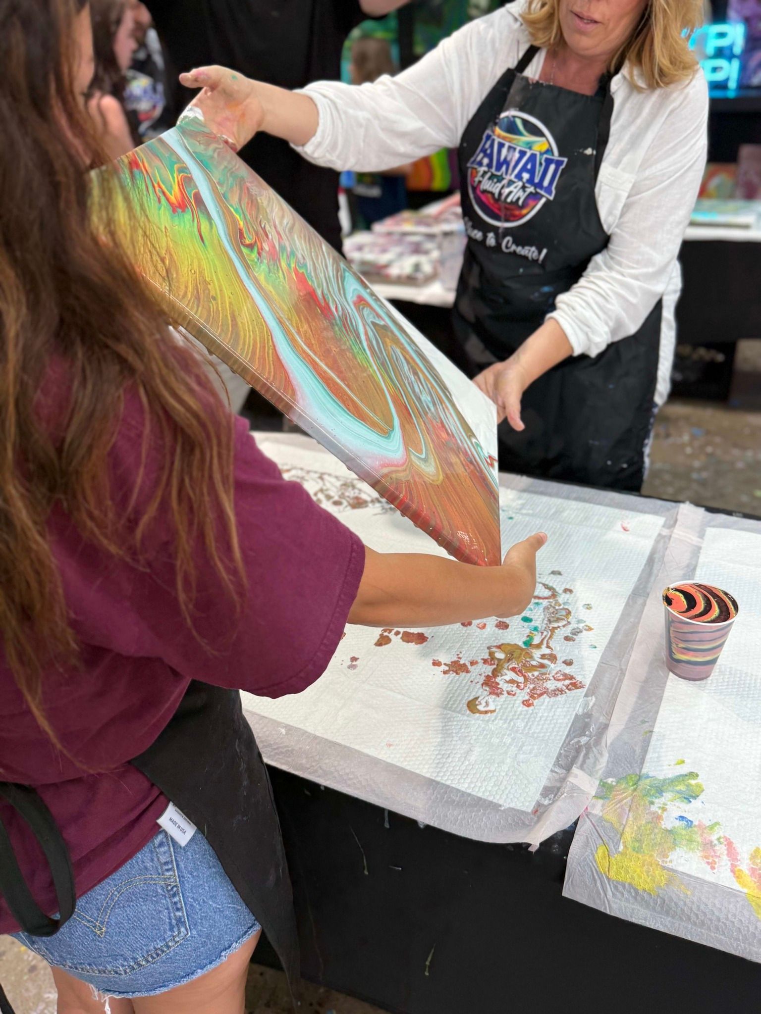 Person in a craft apron helping someone apply glitter to a large iridescent shape at a table