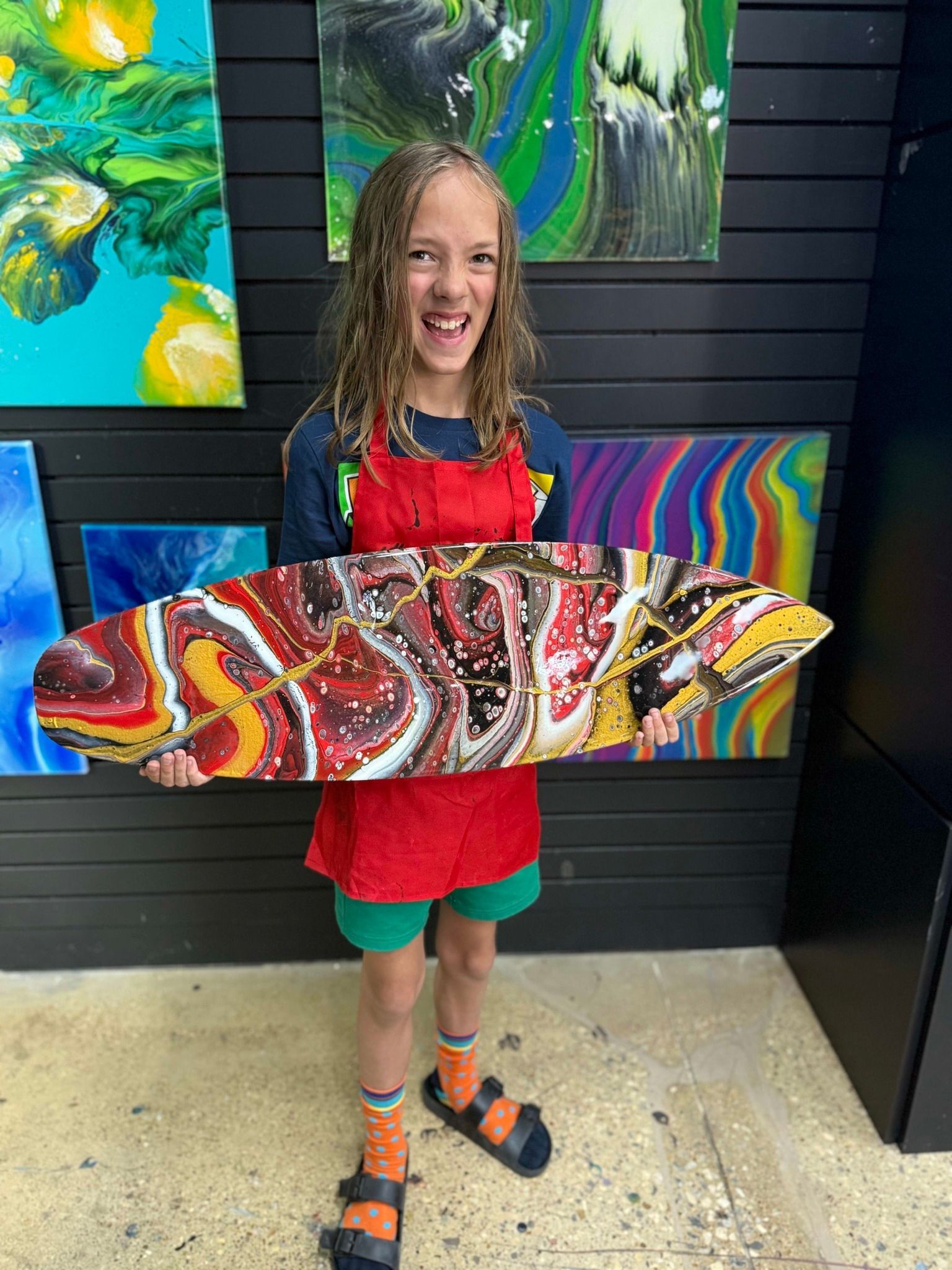 Girl holding a colorful abstract surfboard art piece in front of vibrant paintings