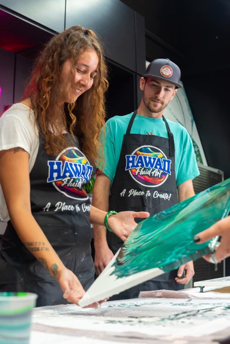 Two people in Hawaii shirts pouring teal paint onto a canvas at a table