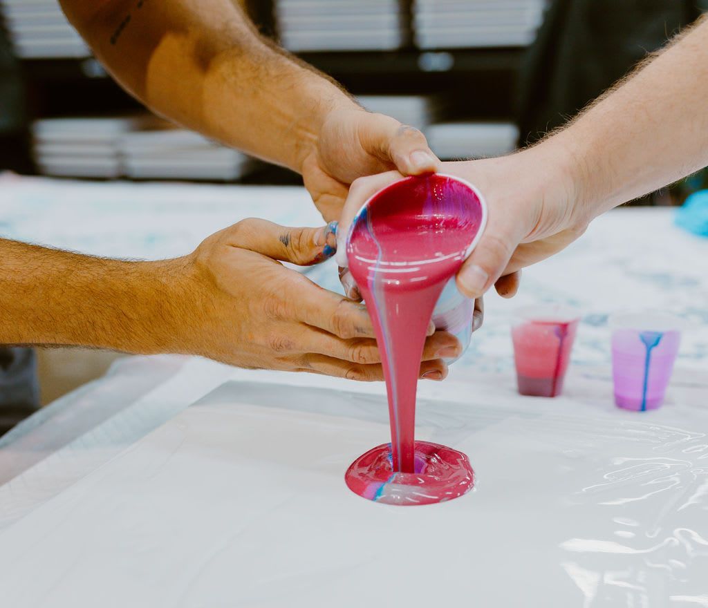 Hands pour bright pink liquid into a small cup on a covered table during a craft activity.