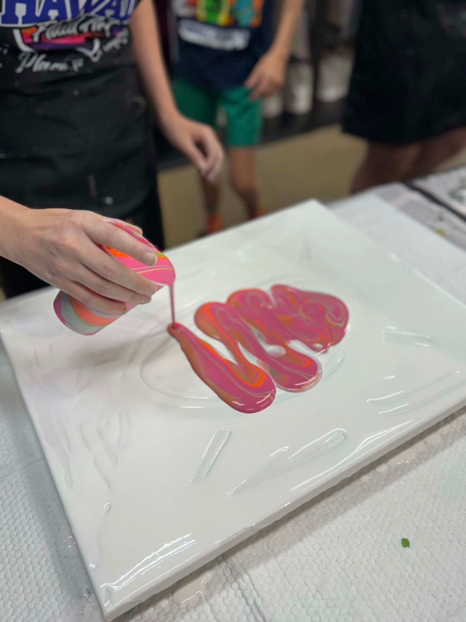 Hand pouring pink paint onto a white canvas at a craft table