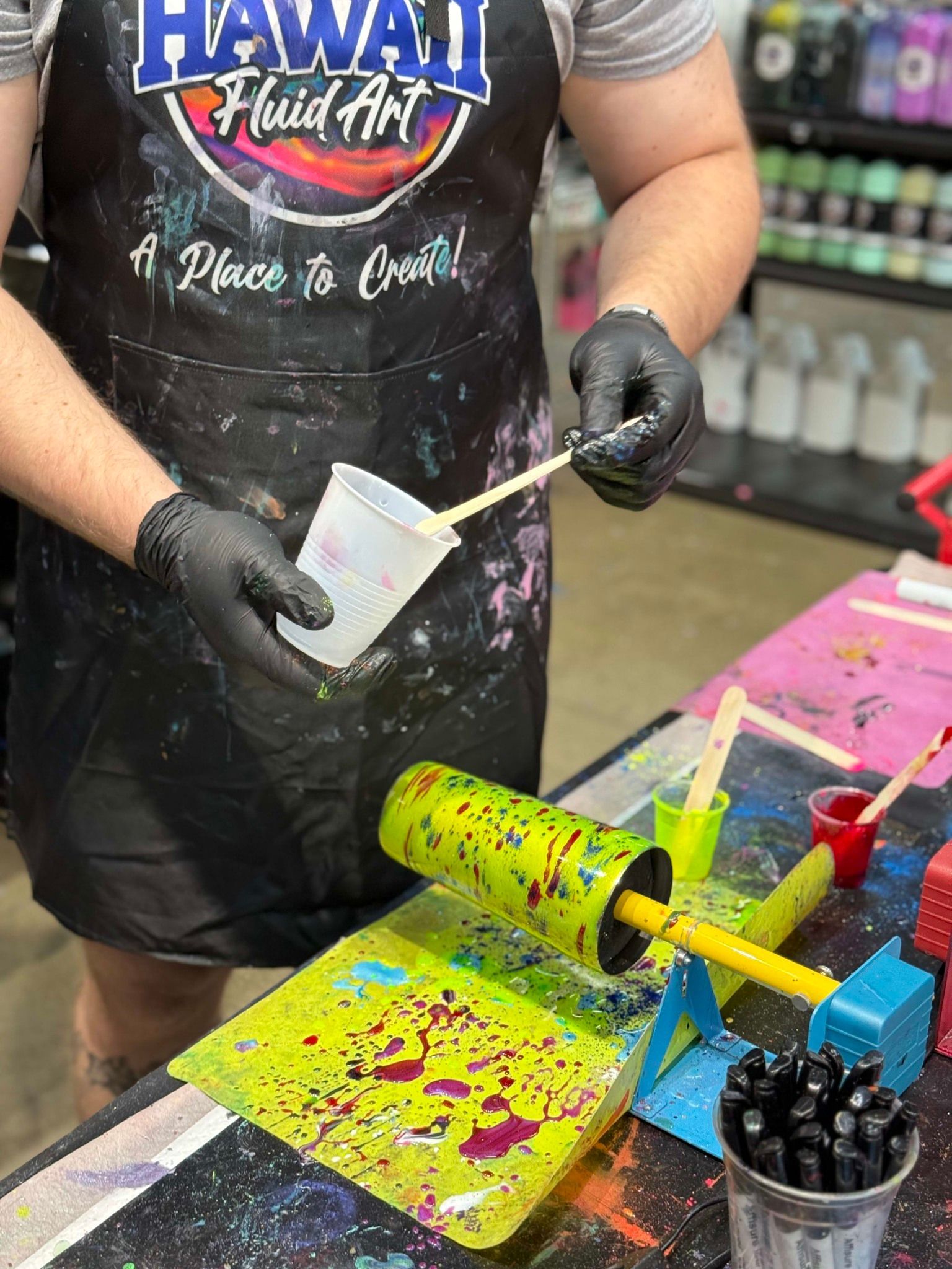 Person wearing a Hawaii Hula t-shirt pouring liquid into a cup at a colorful tie-dye station