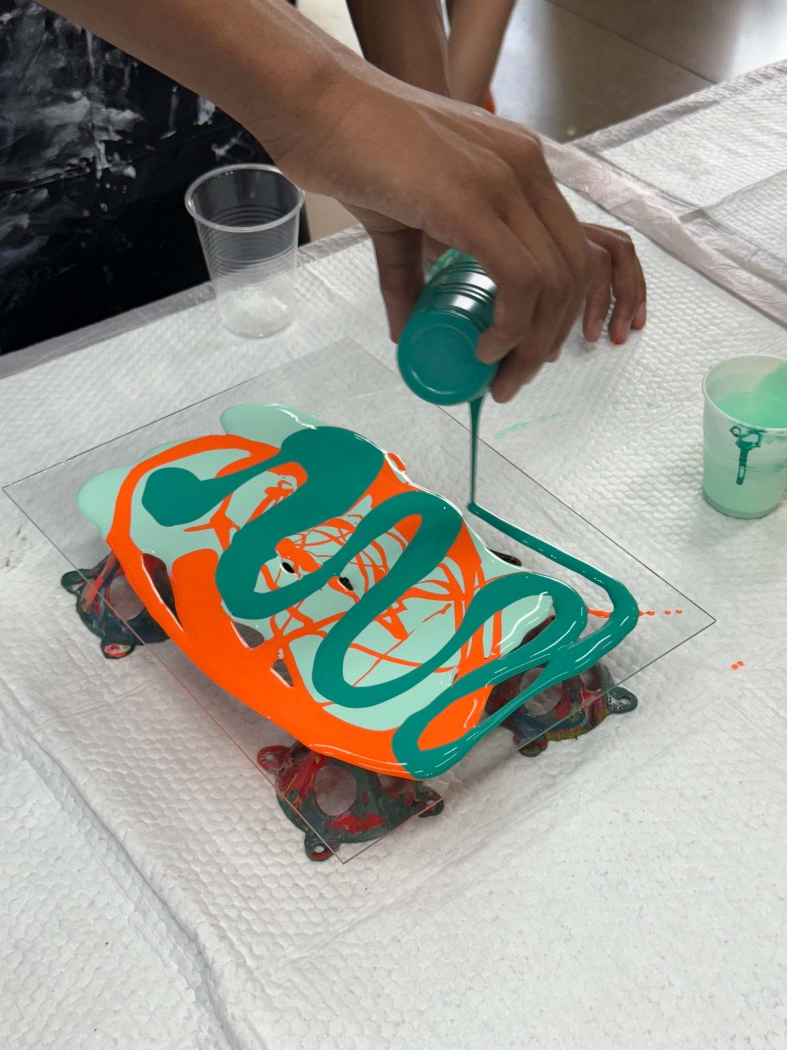 Hand pouring green paint over an orange and teal tray on a table, with cups nearby