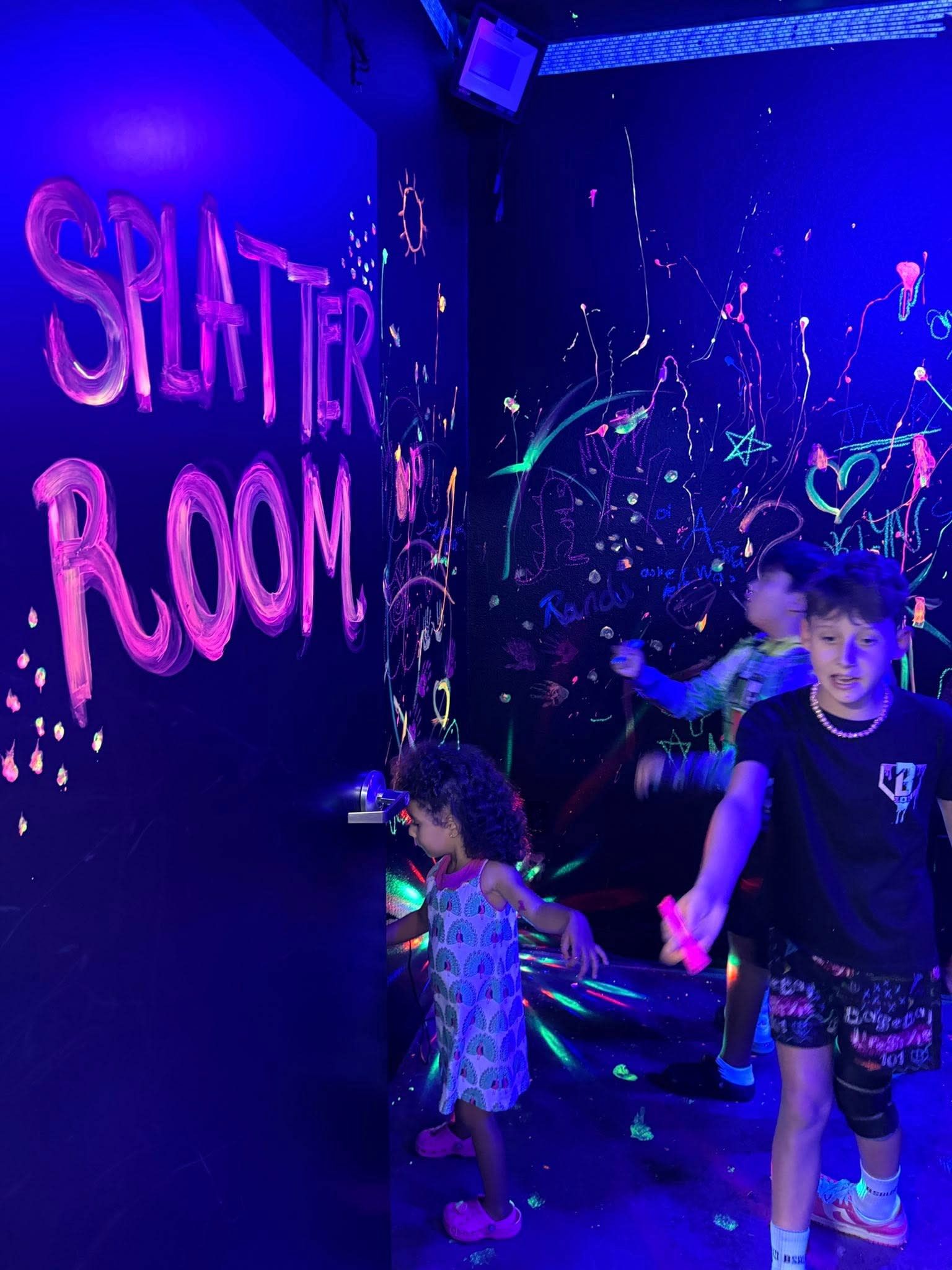 Children in a neon-lit splatter room, playing near glowing paint splashes and a sign reading “SPLATTER ROOM”
