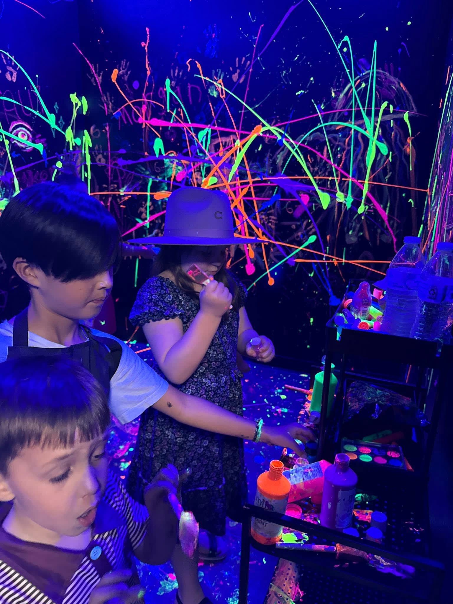 Children painting with neon glow paint at a dark art station, surrounded by colorful splashes and lights