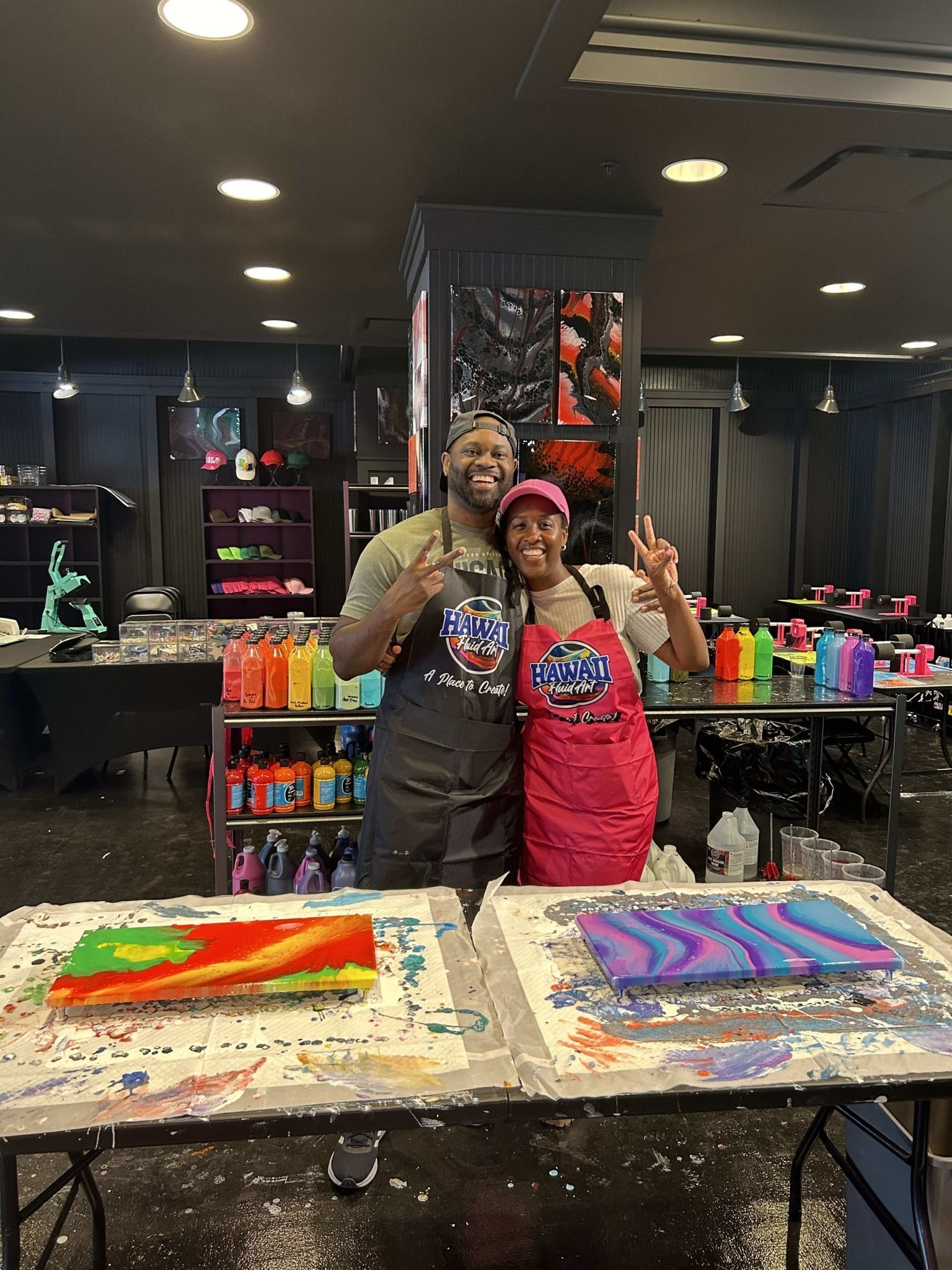 Two people posing in an art studio beside colorful abstract paintings on tables