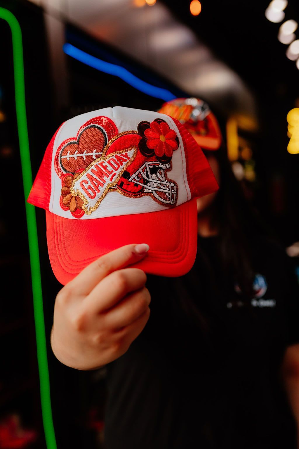 Hand holding a red baseball cap with heart and flower patches in a dim, colorful setting