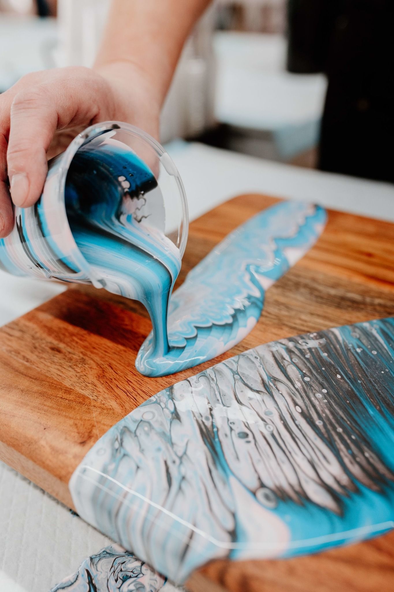 Hands pouring blue resin over a wooden board, creating a glossy abstract finish.