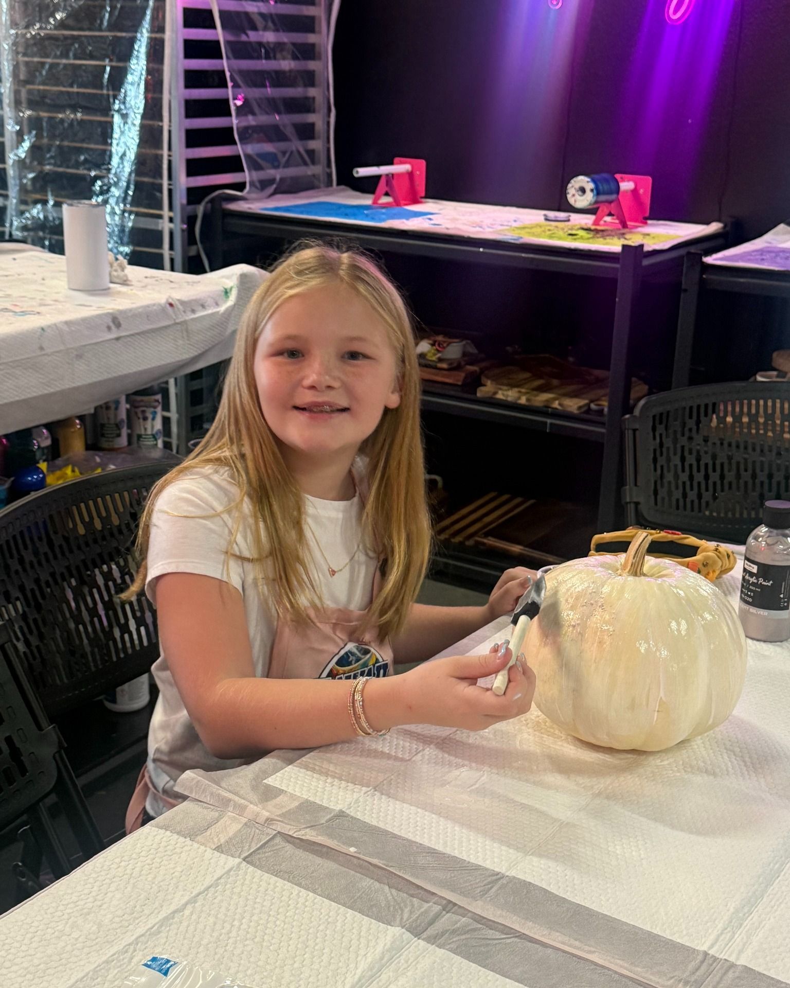 Child smiling at a table, painting a white pumpkin in a decorated indoor workshop.