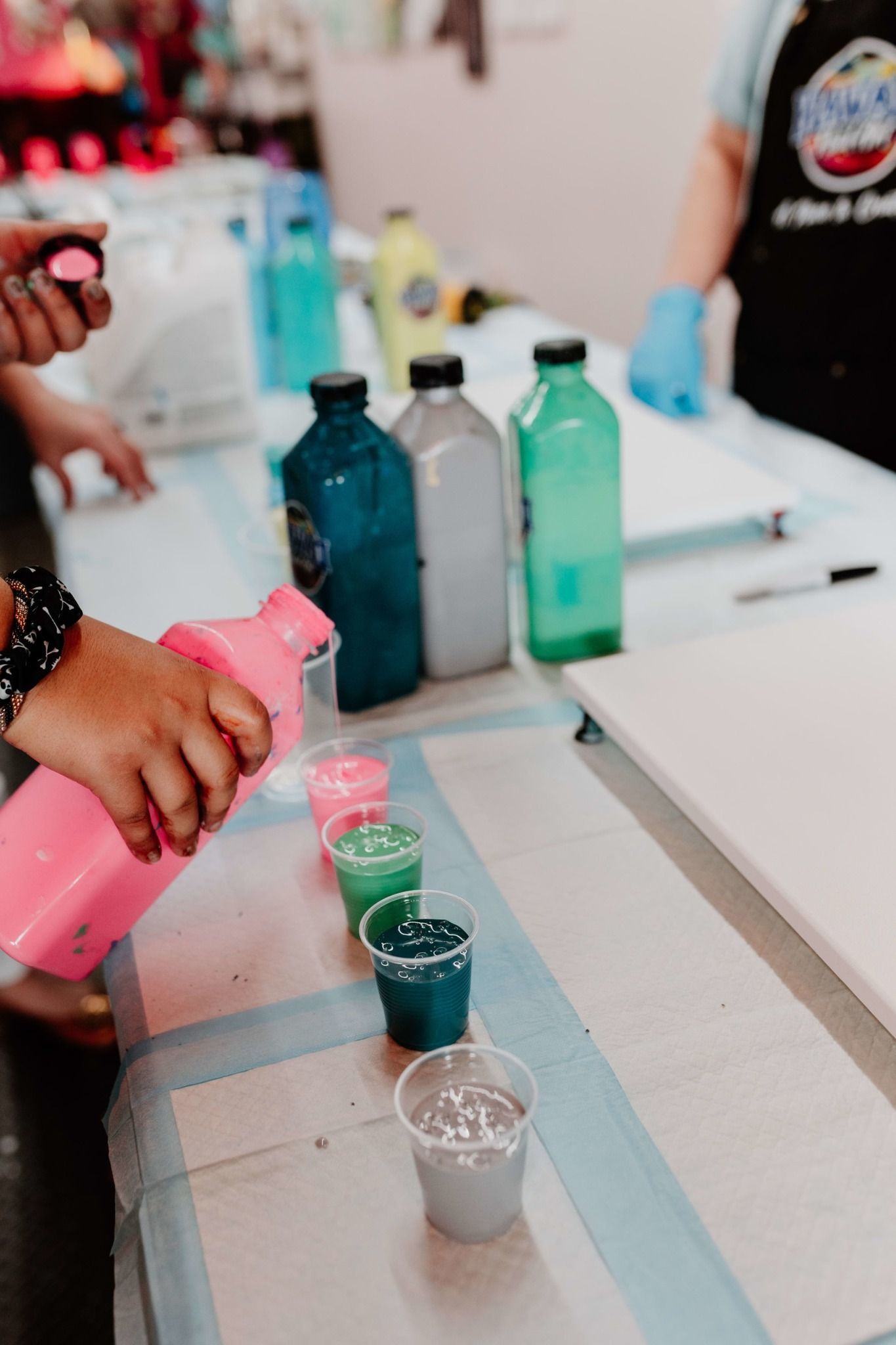 Hands pouring colorful liquids into small cups beside bottled drinks on a table