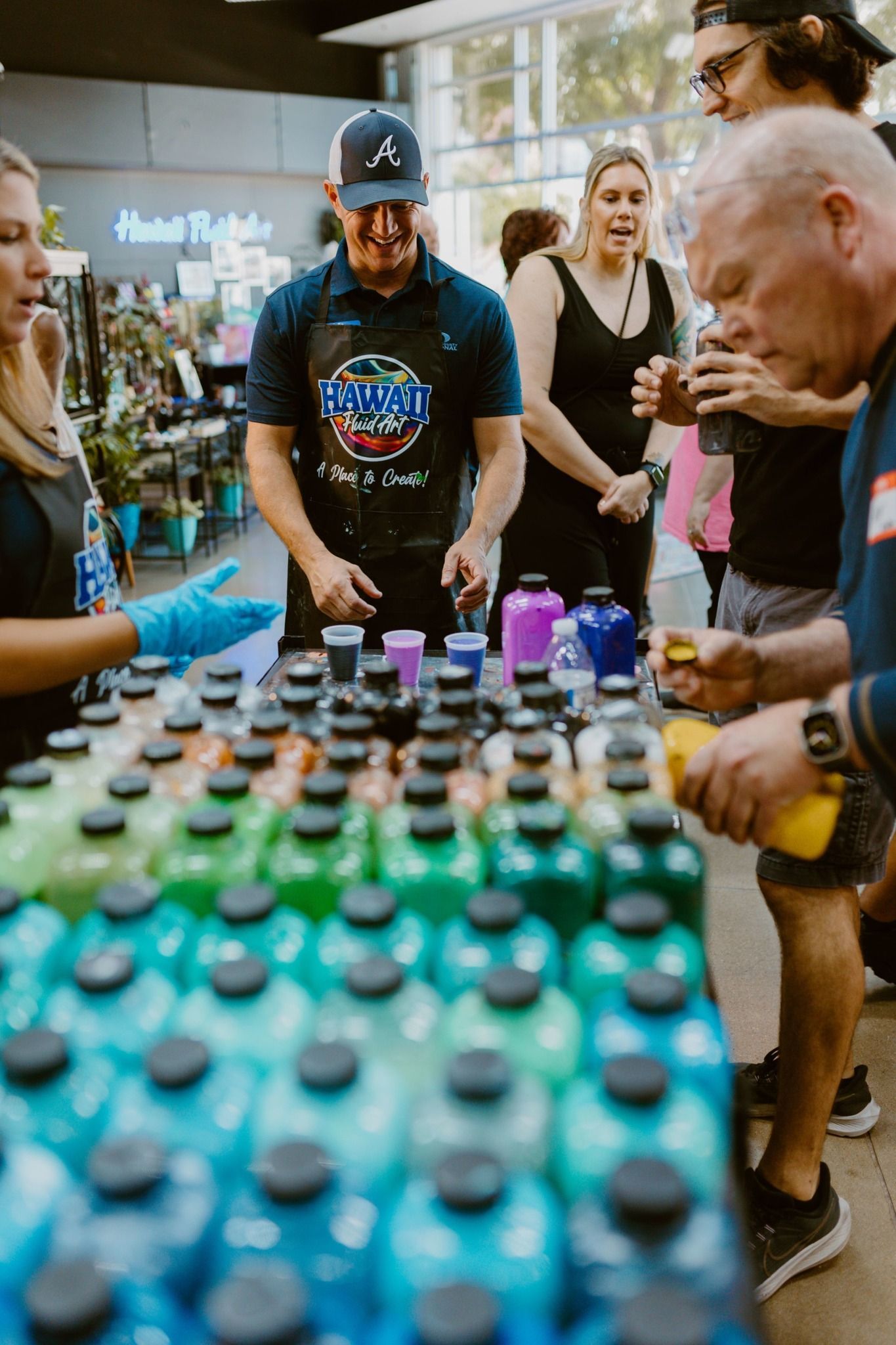 People sampling colorful bottled drinks at a market stall, with a vendor and crowd around the table