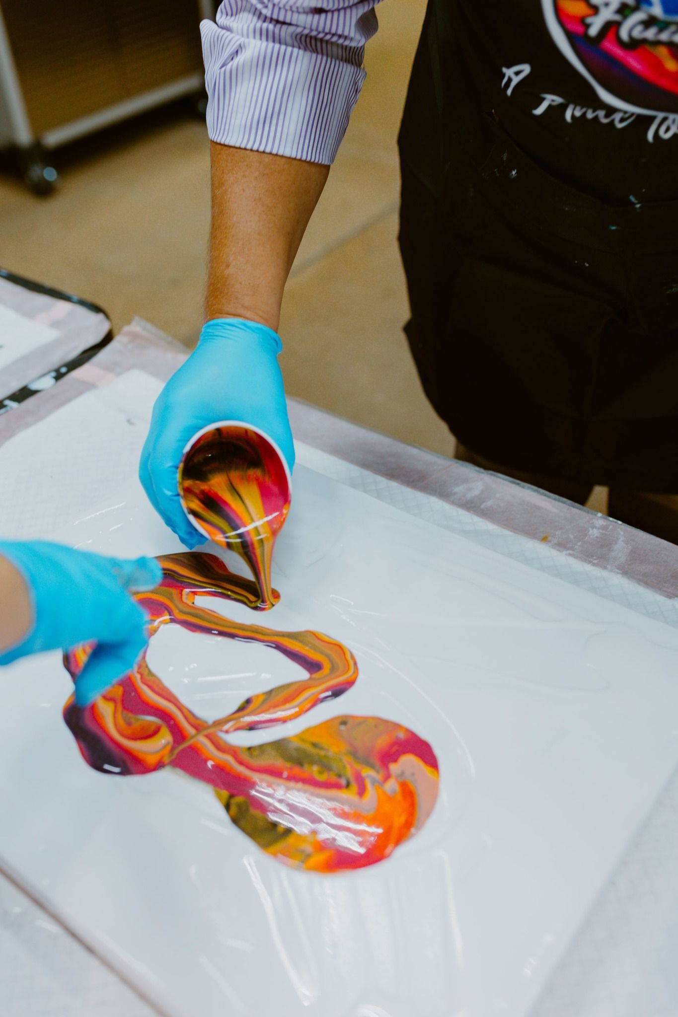 Gloved hands pouring colorful liquid paint onto a white canvas