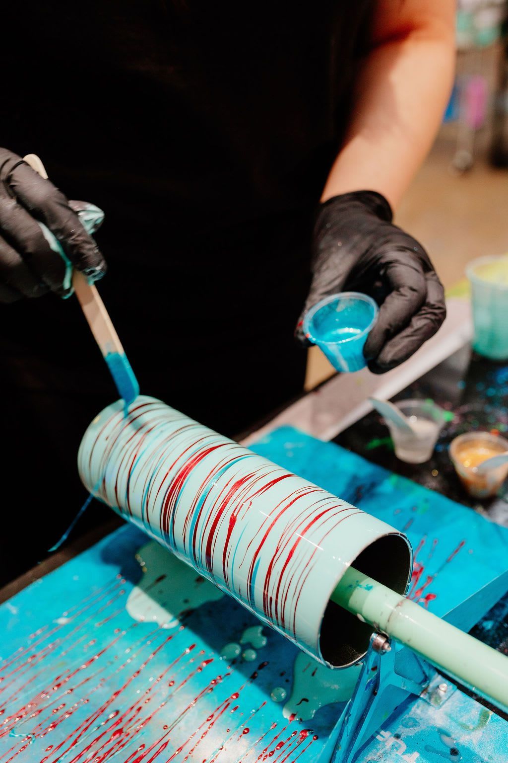 Hands pour blue liquid onto a rotating white cylinder with red stripes in a studio workspace