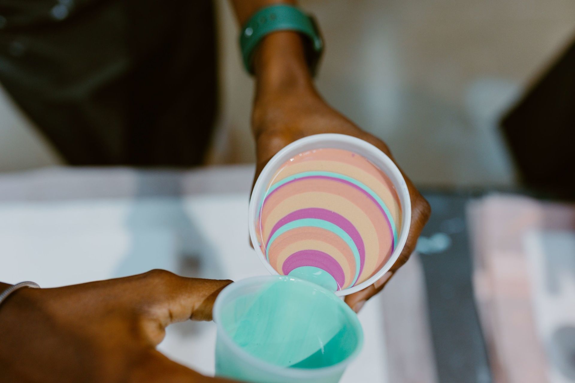 Hands pouring pastel liquid from striped cups into a turquoise cup