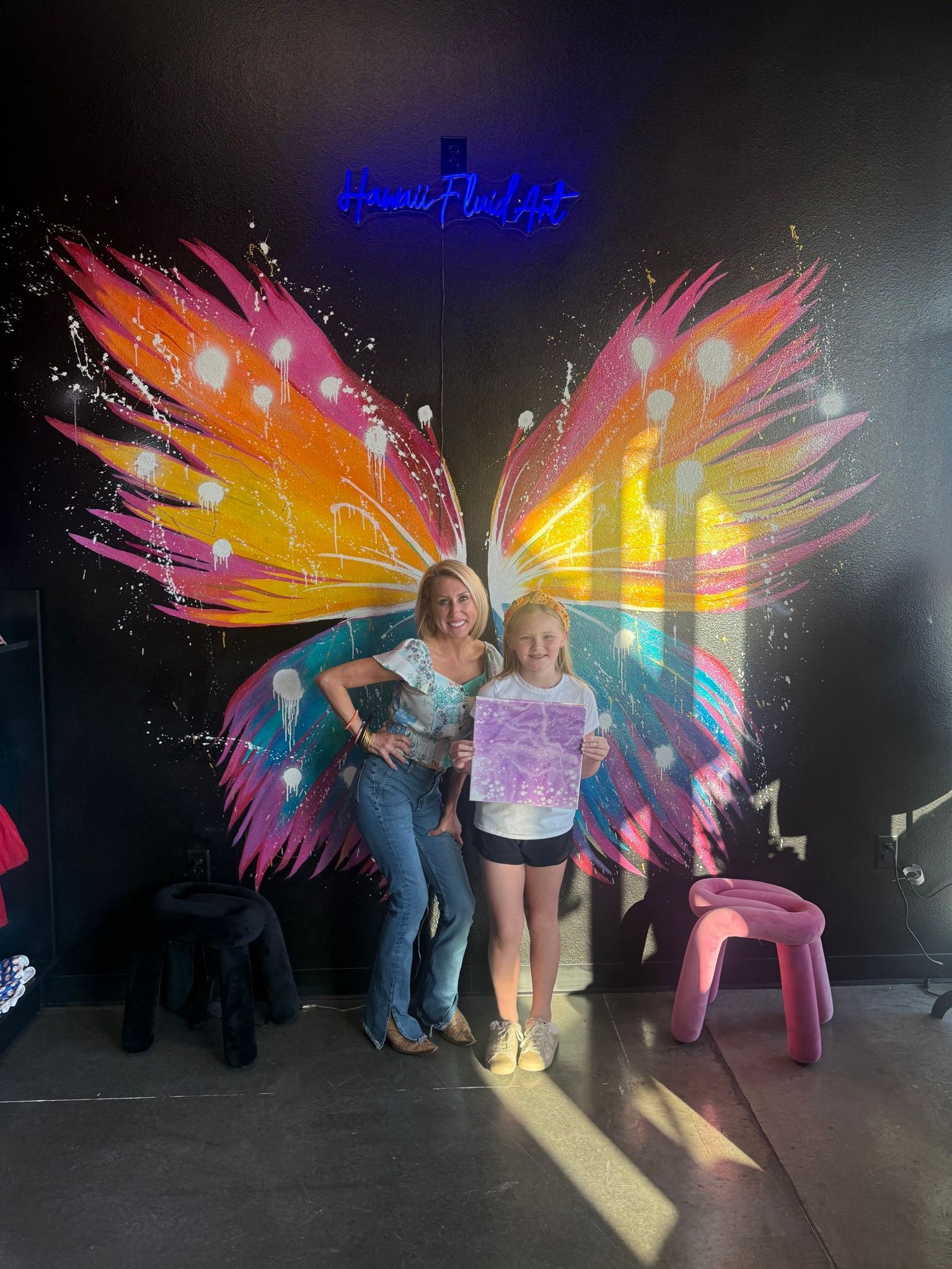 Two people posing in front of colorful butterfly wings mural with neon lights and chairs nearby