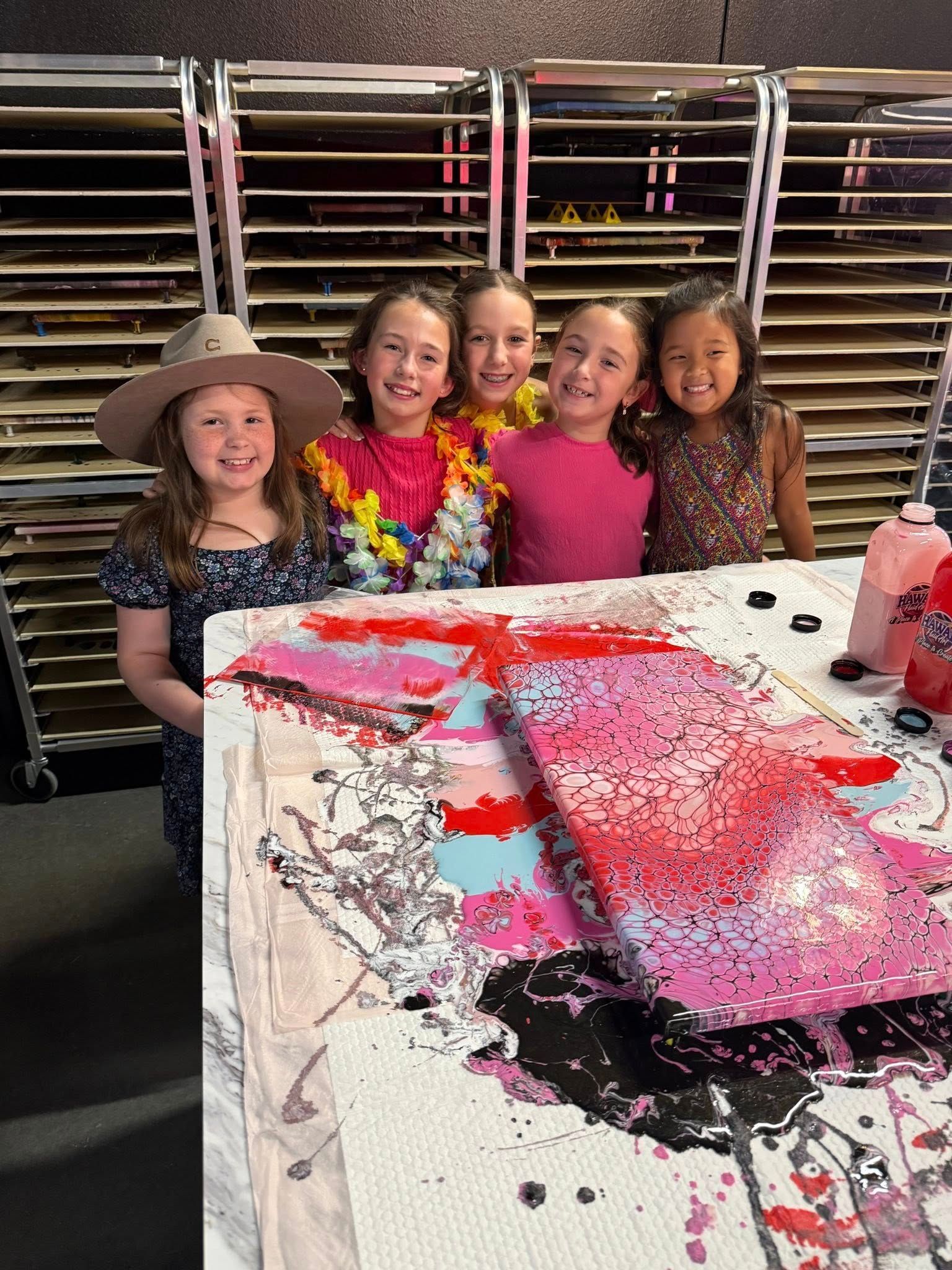 Five smiling children stand behind a paint-splattered table with a colorful art project.