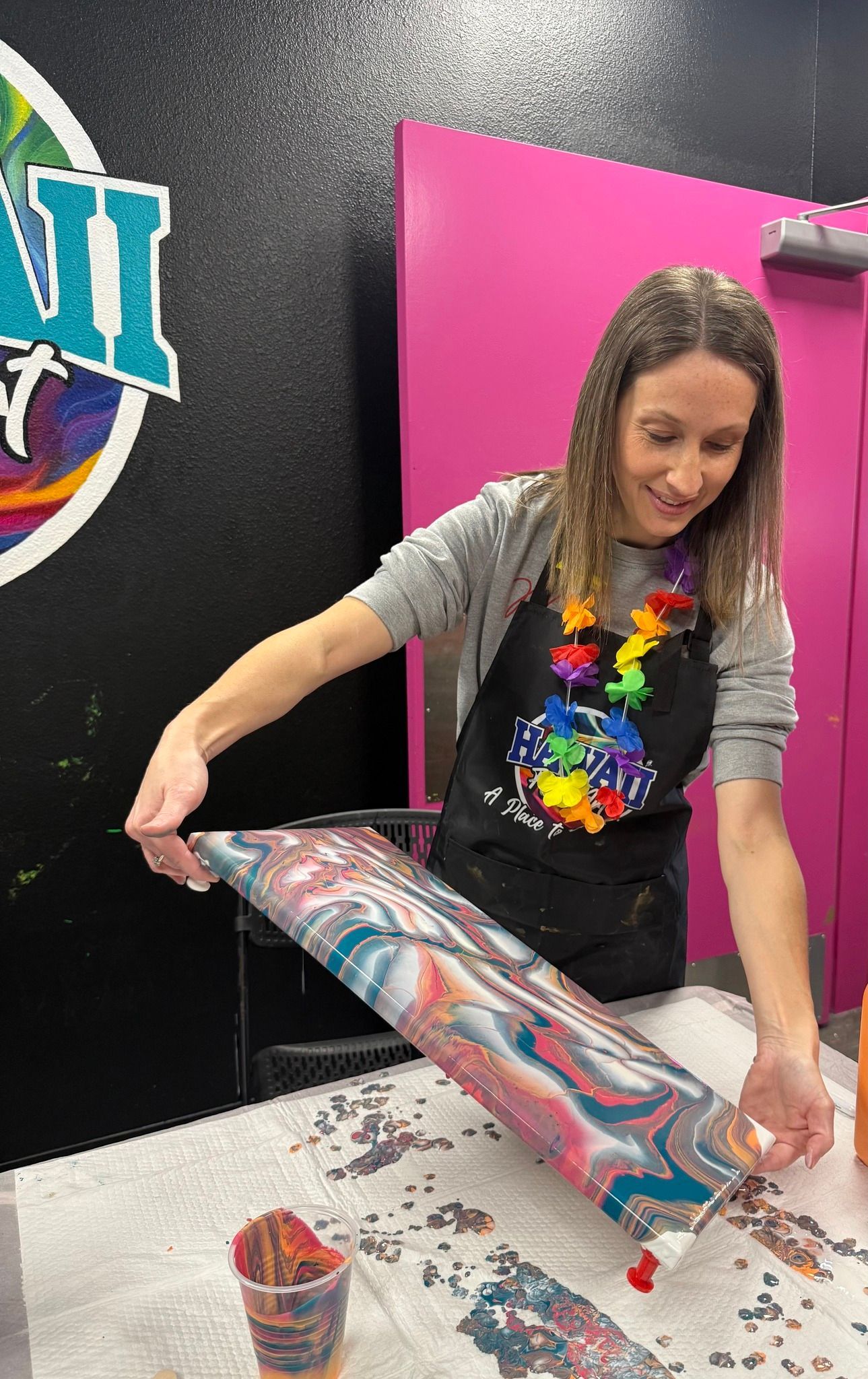 Woman pouring colorful paint onto a tilted canvas in a studio with a pink door and black wall