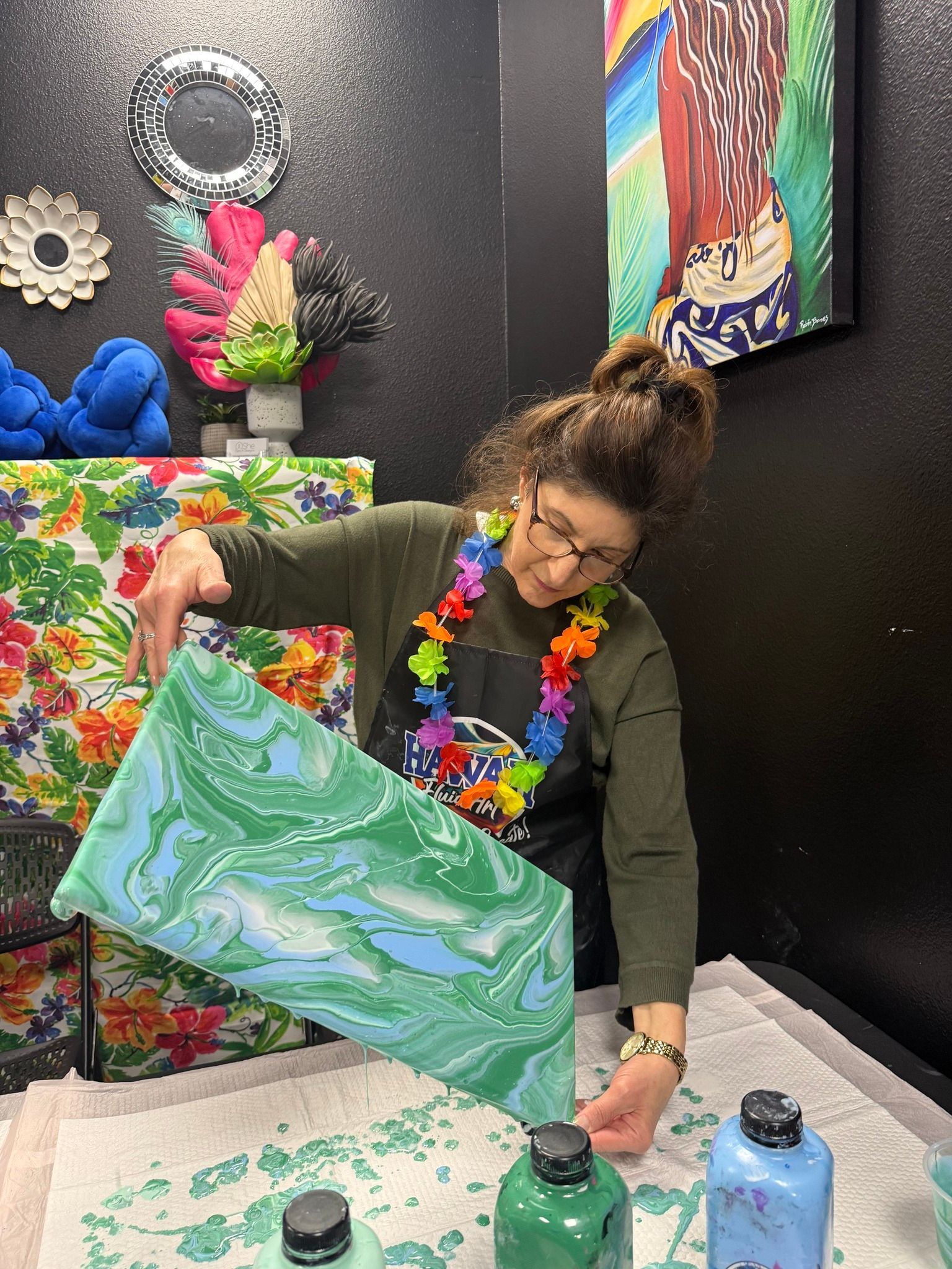 Person painting a green canvas at a table with paint bottles in a colorful art studio.