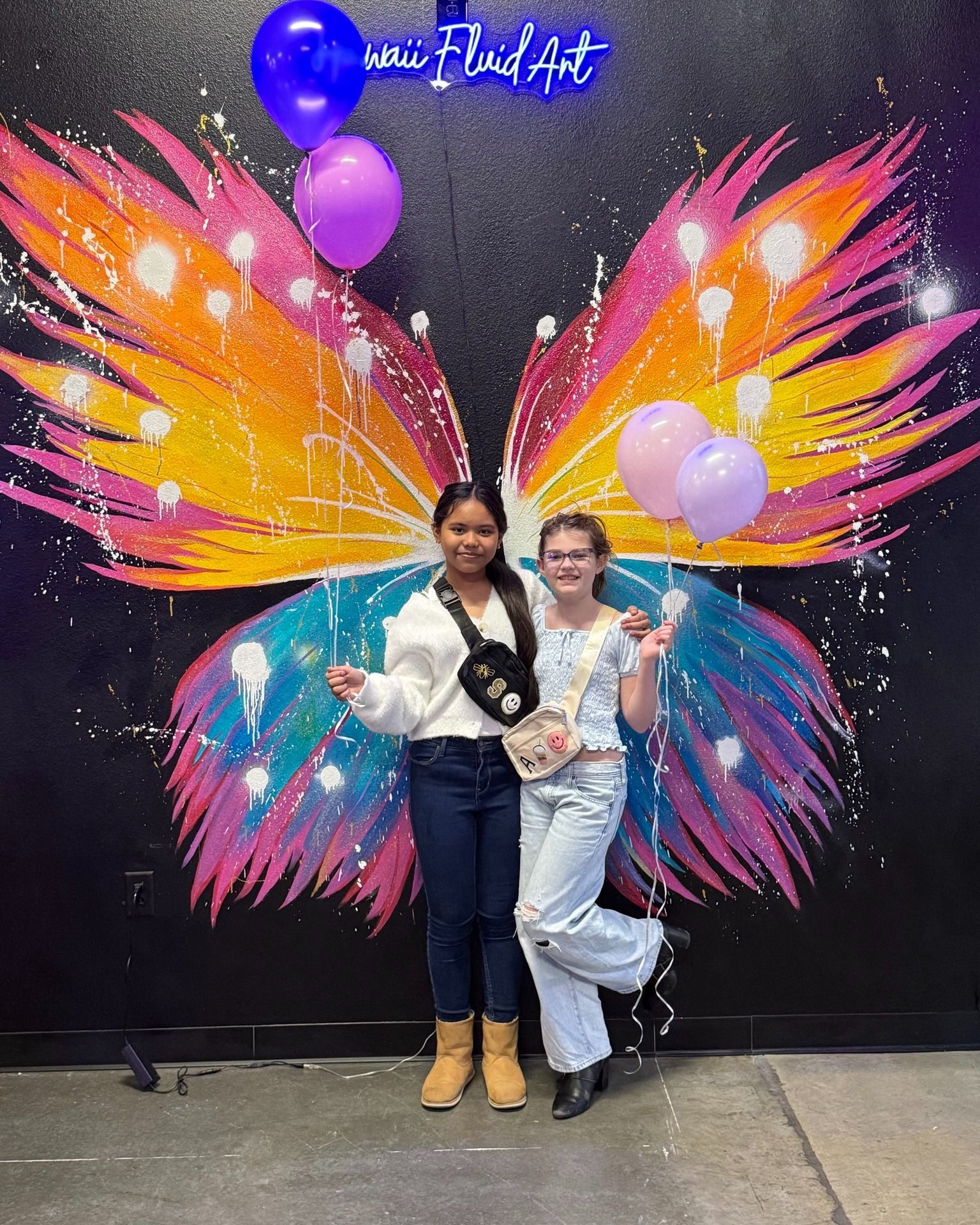 Two people posing in front of a colorful butterfly wing mural with balloons and a neon sign.