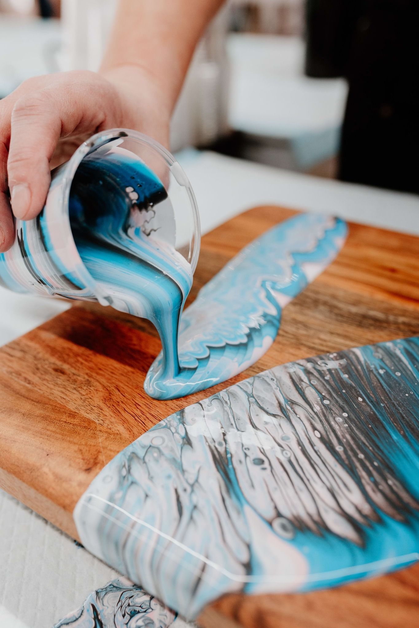 Hands pouring blue resin onto a wooden board, creating a glossy abstract pattern.