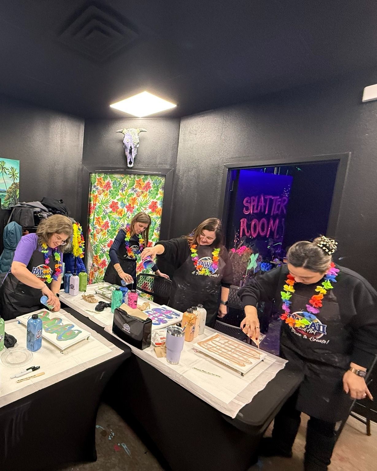 Four people in colorful shirts work at tables in a dark room with neon decor and a glowing “SHUTTLE ROOM” sign.
