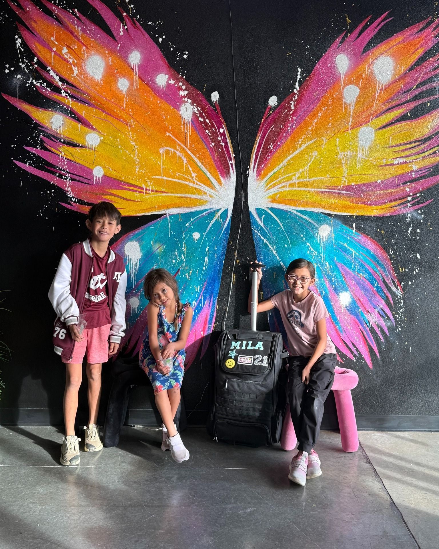 Three children posing in front of a colorful butterfly mural with a black display stand.
