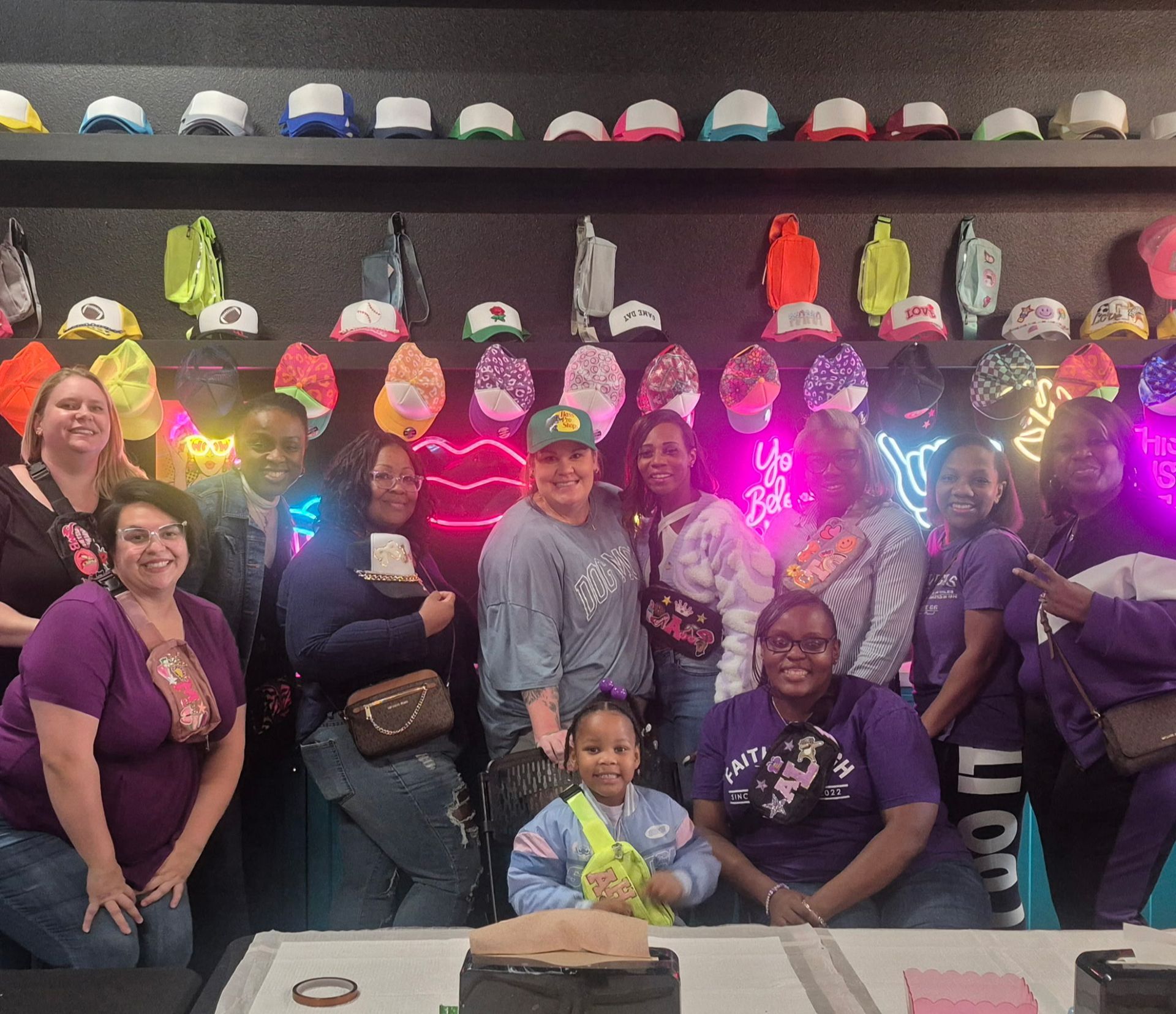 Group posing indoors in front of a wall of colorful hats and glowing lights, smiling at the camera.