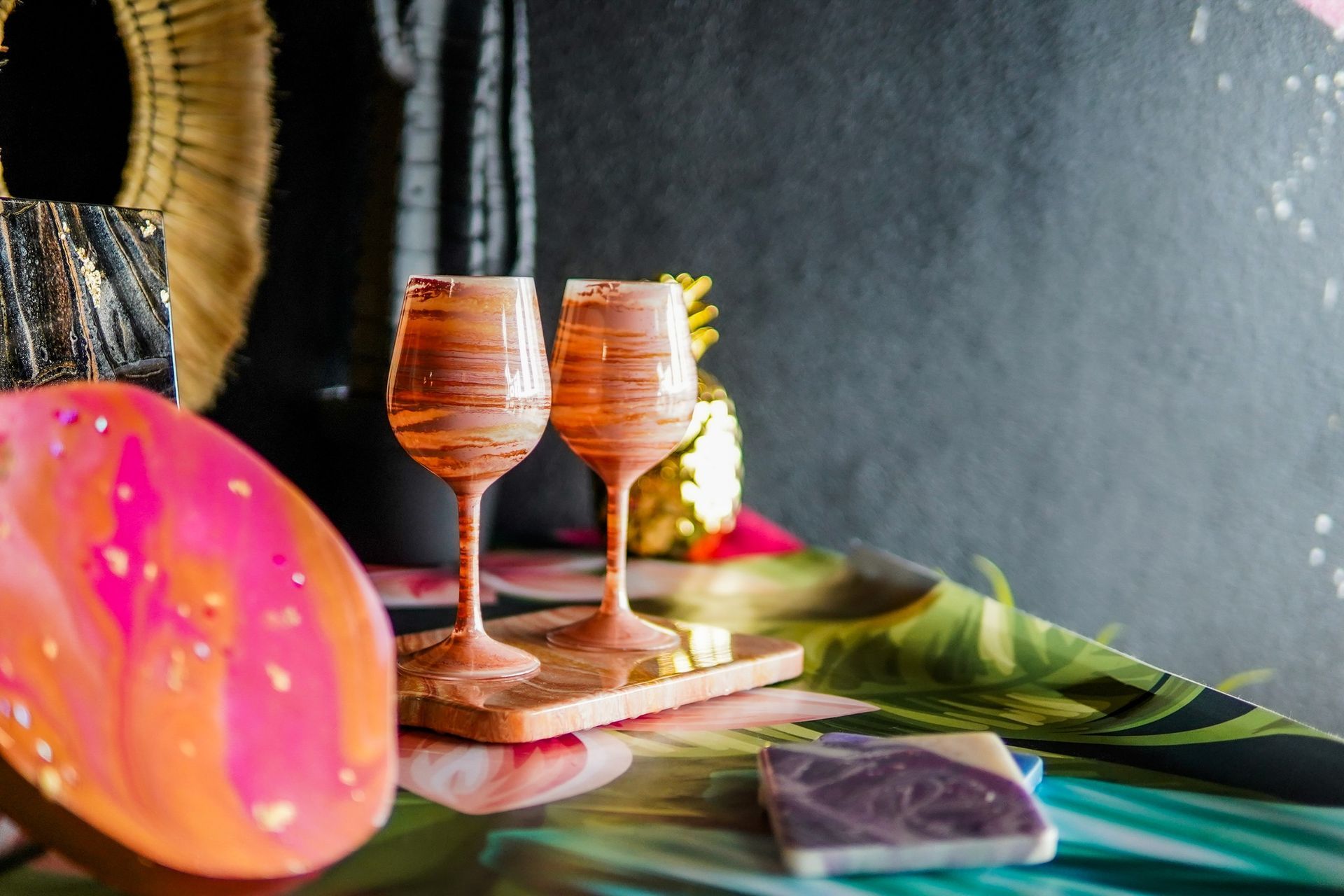 Two orange goblets on a wooden tray beside a pink citrus slice and green palm leaf on a dark table