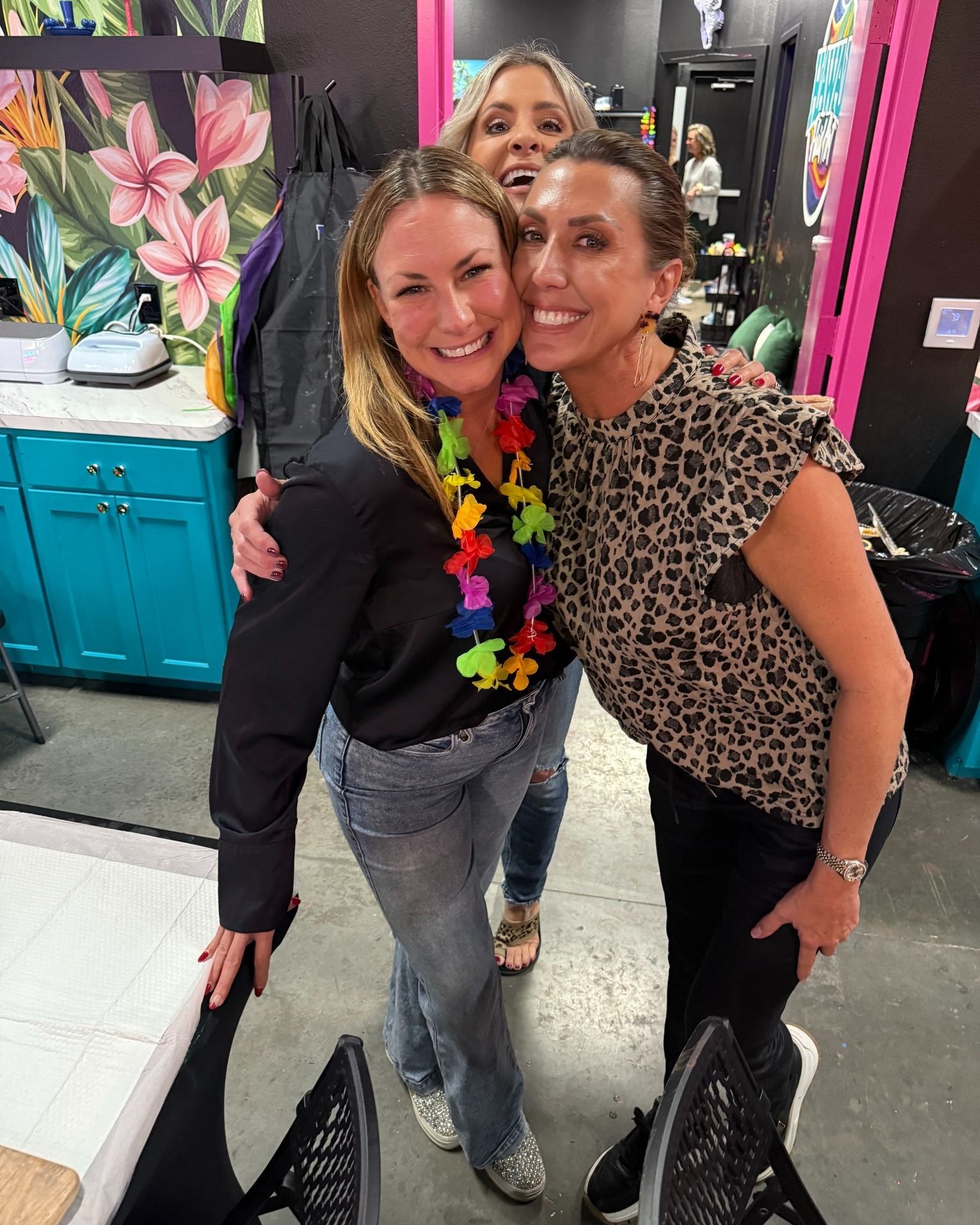 Three smiling friends posing in a salon with colorful cabinets and a floral painting behind them.