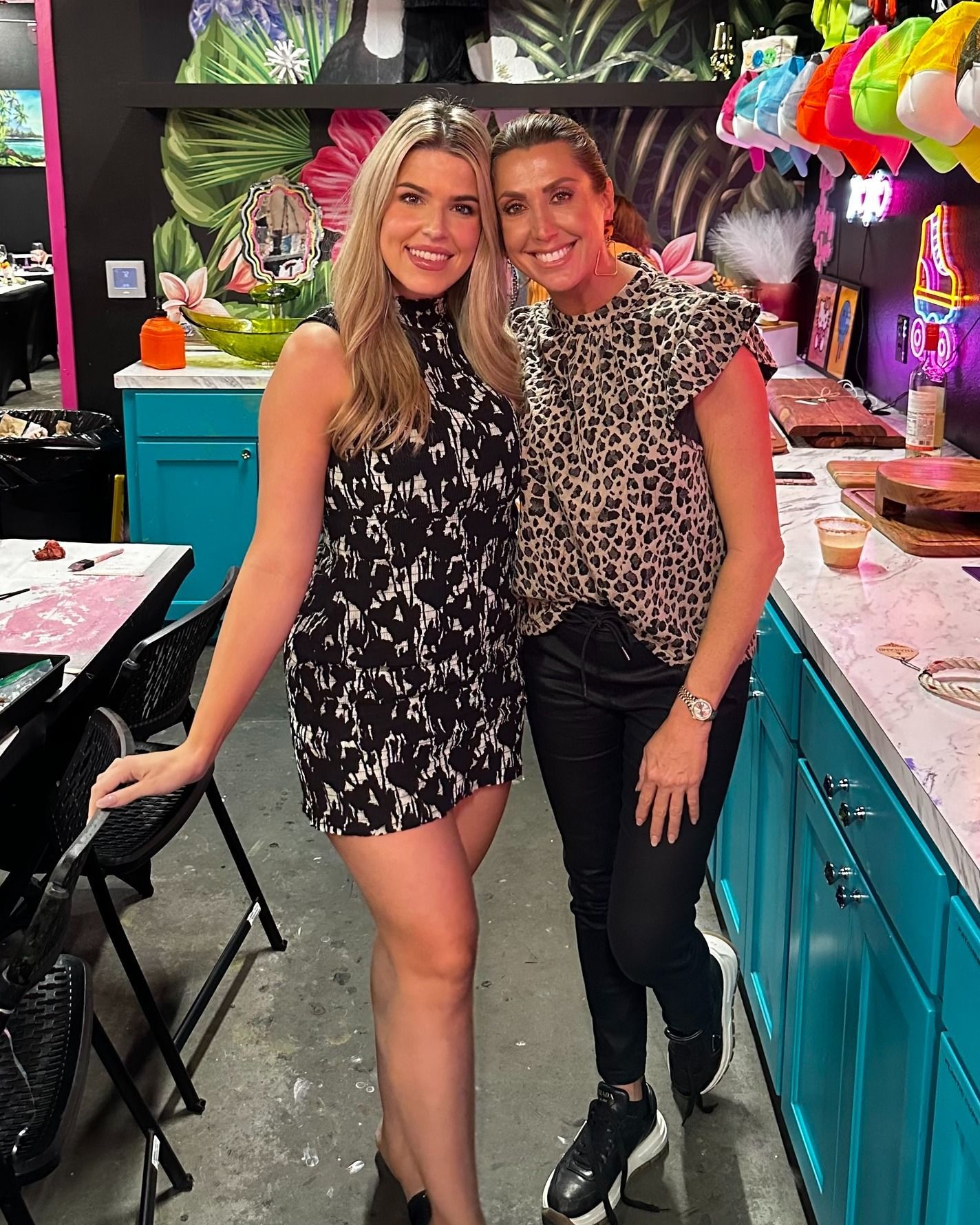 Two women smiling and posing in a colorful salon with teal cabinets and bright decor