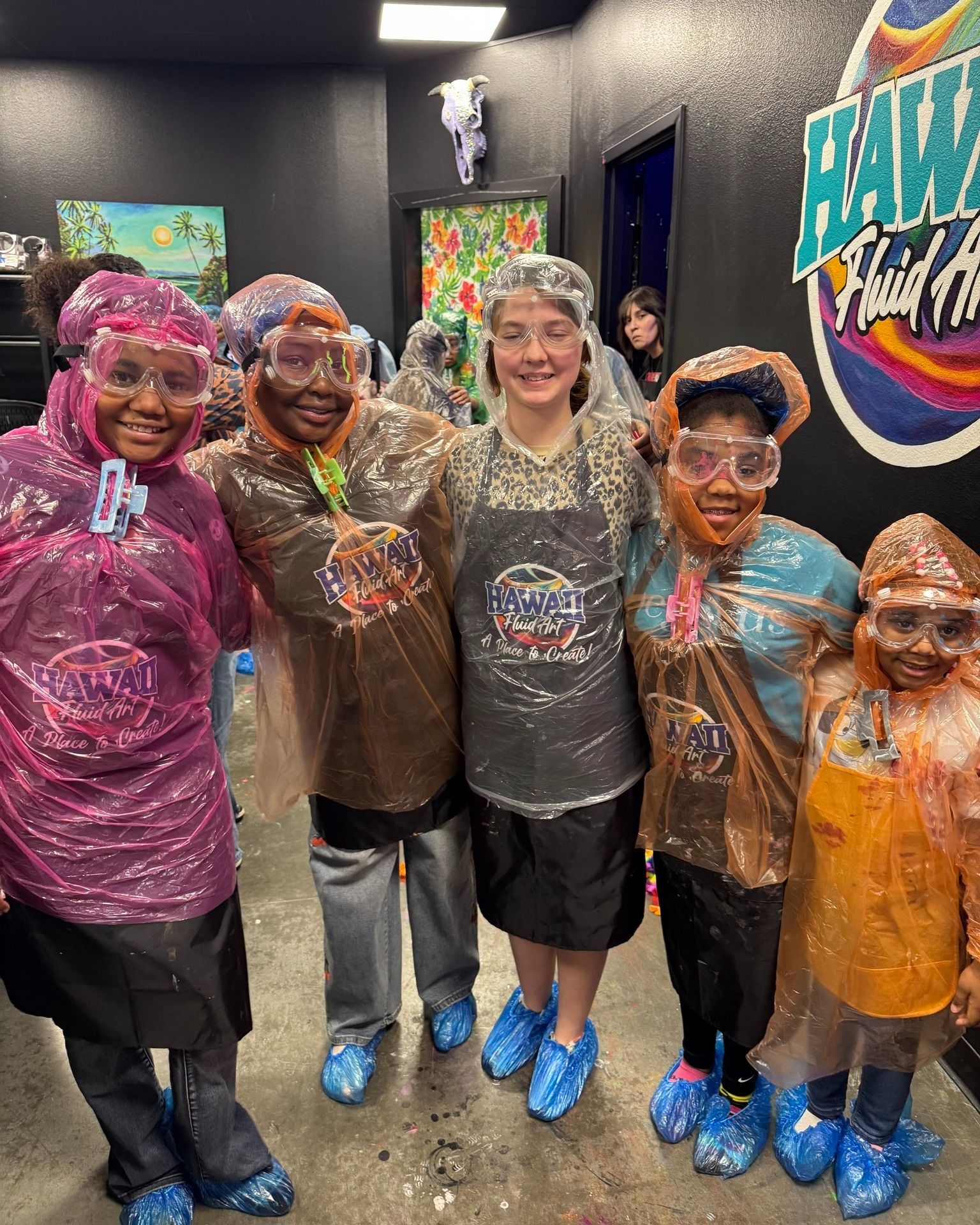Five people in rain ponchos and blue shoe covers smiling indoors at an art studio wall mural.