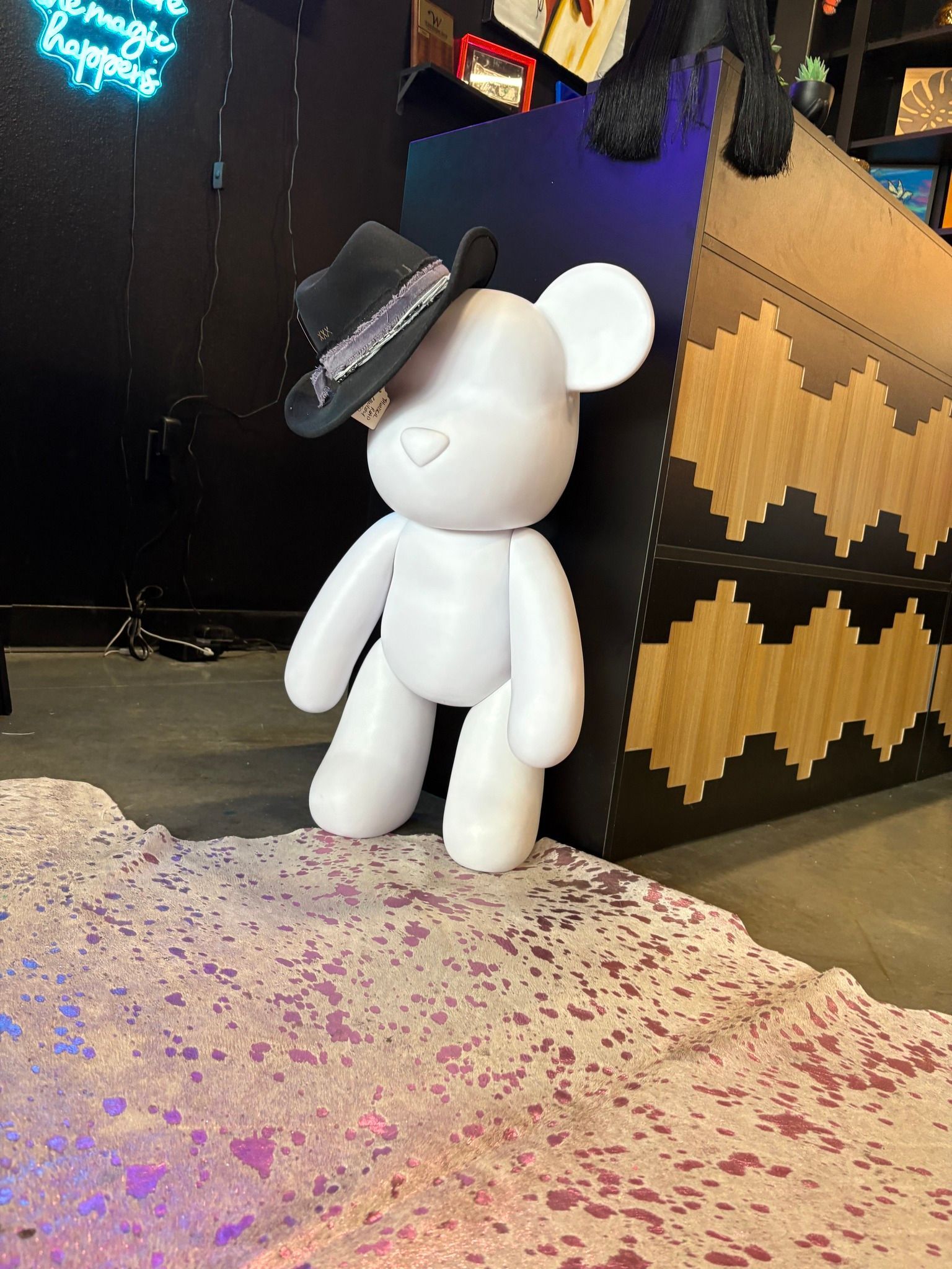 White bear-like figure in a black hat standing on a speckled floor beside a geometric black-and-gold wall.