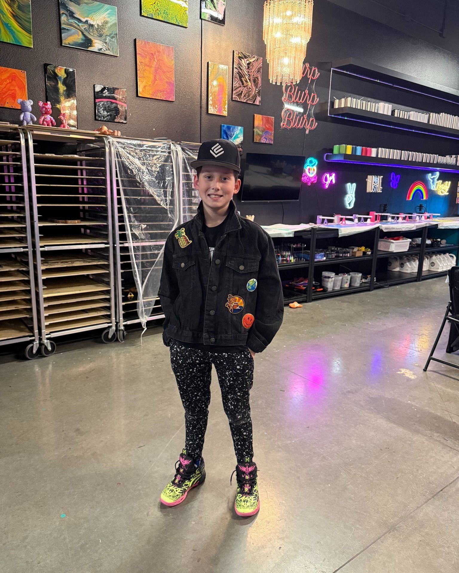 Person standing in a bowling alley, wearing a black jacket, patterned pants, and neon shoes.