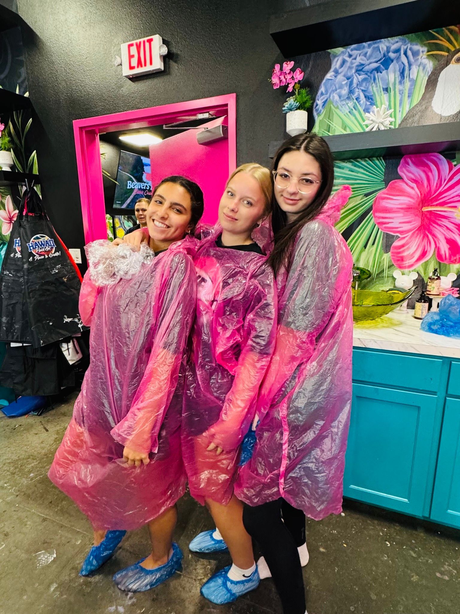 Three smiling people in pink rain ponchos pose in a colorful shop or studio.