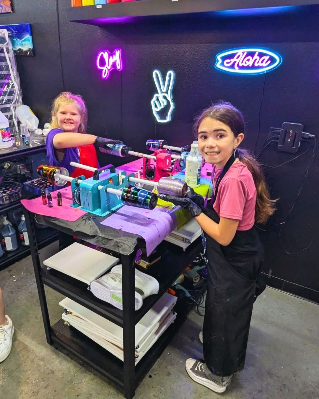 Two kids at a colorful neon-lit craft table with bottles, tools, and a peace-sign wall sign