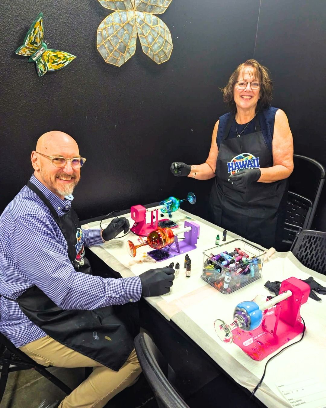 Two people at a craft table with colorful small machines and tools, smiling in a workshop setting