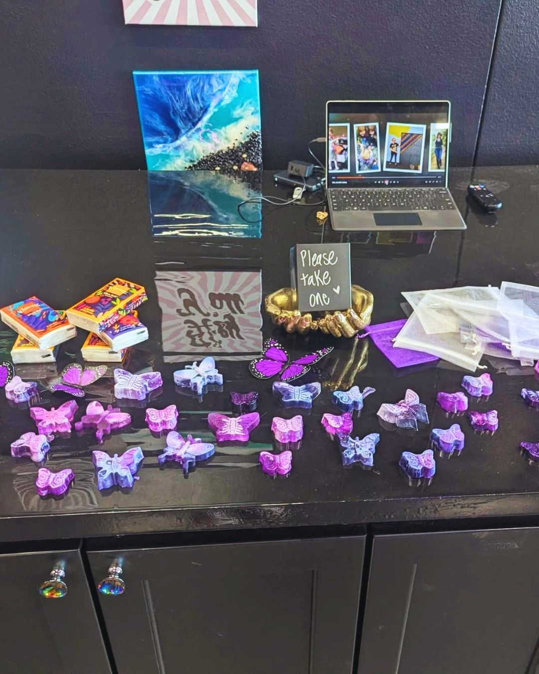 Table display with purple butterfly crafts, art prints, a laptop, and small packaged items on a black surface