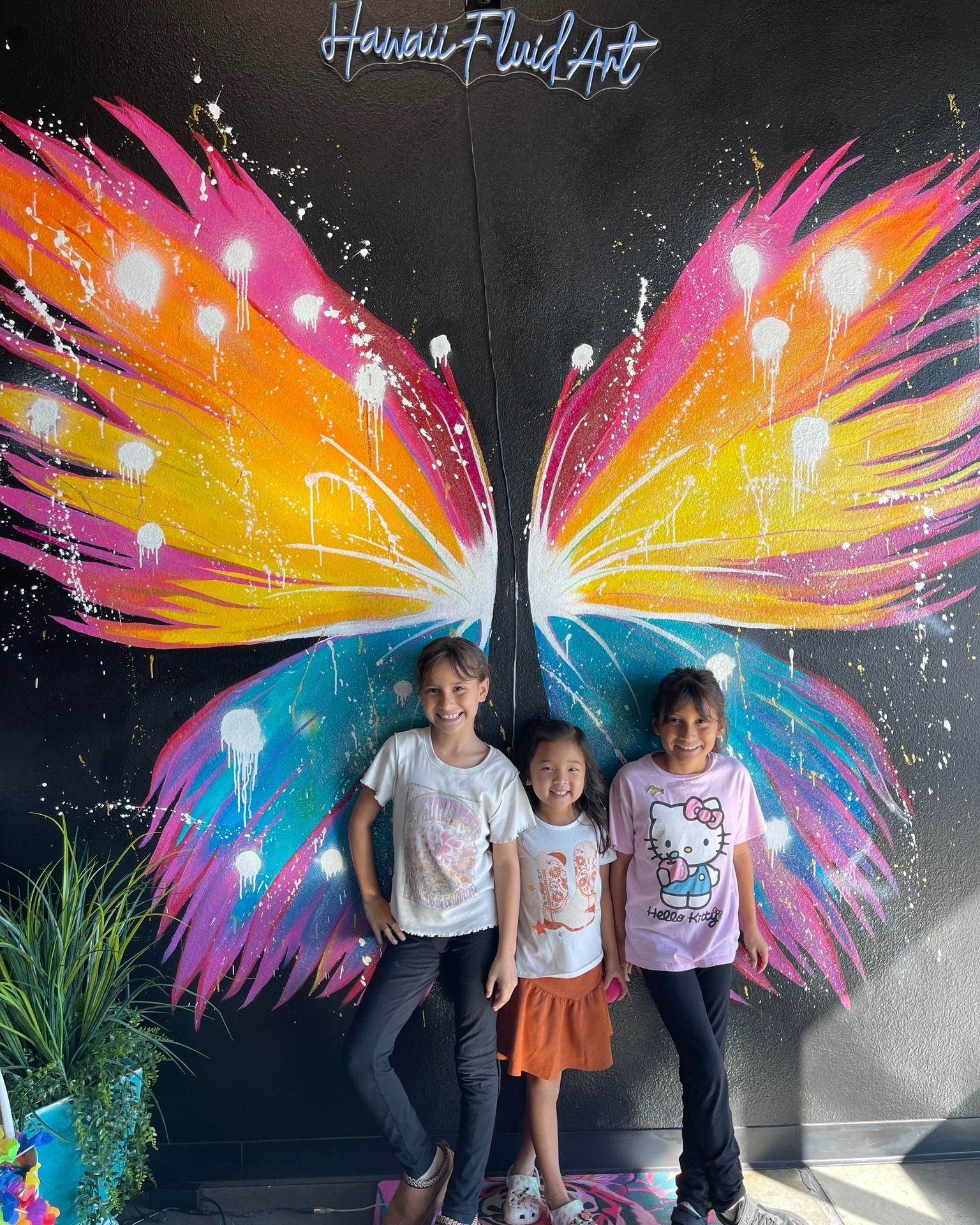 Three children posing in front of a colorful butterfly mural with bright pink, orange, and blue wings
