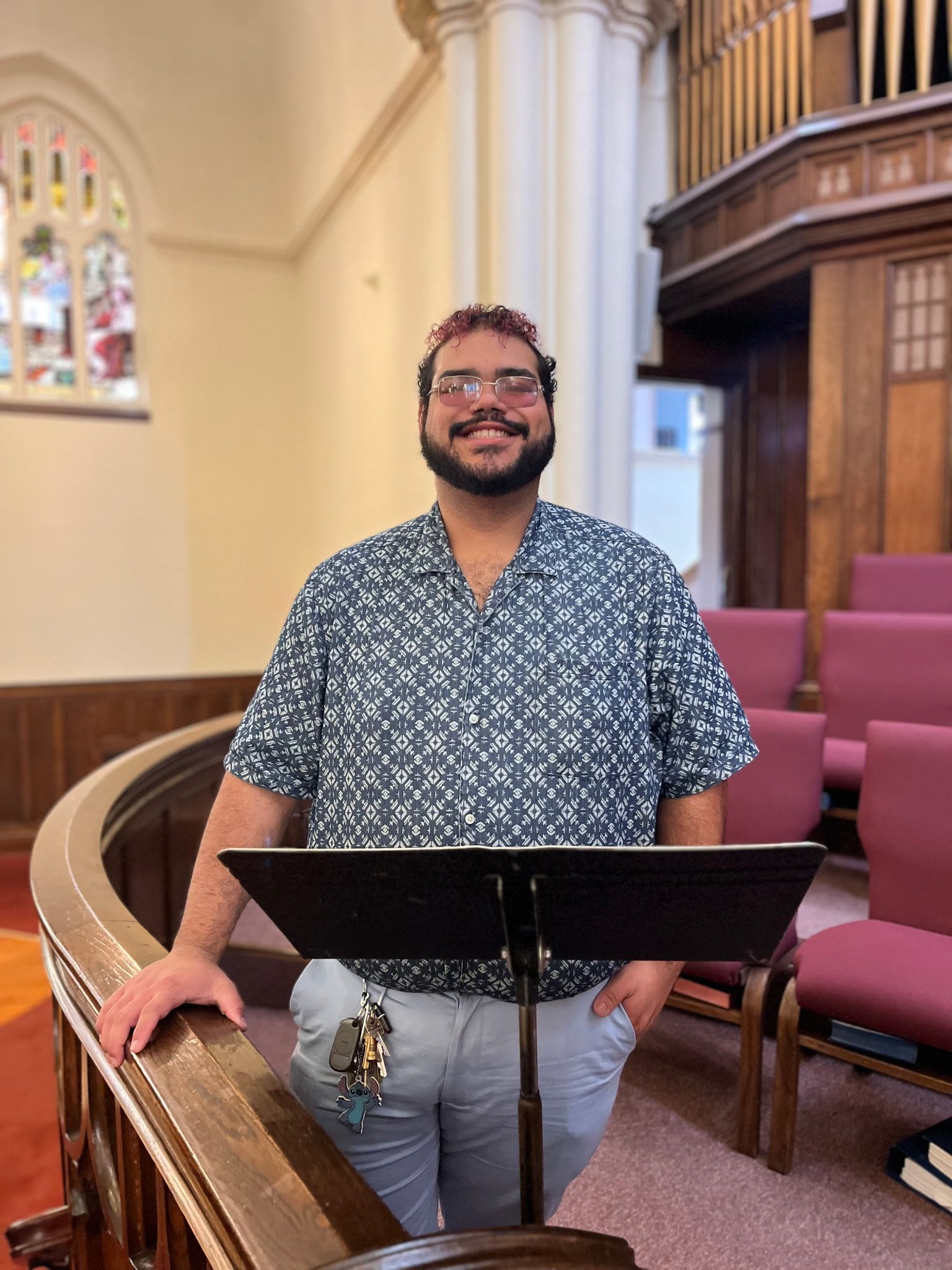 Man standing at a church lectern, smiling, with stained glass and purple chairs in the background