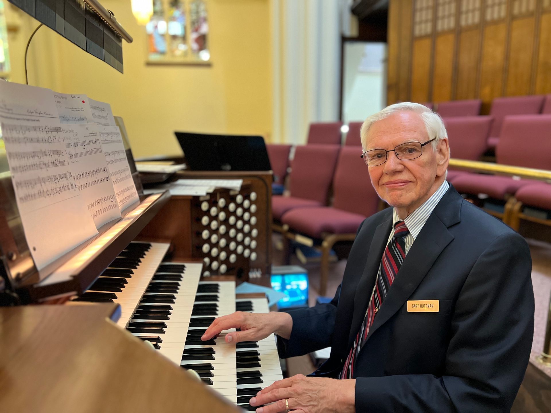 Man playing a church organ beside sheet music, seated in a sanctuary with pink pews and stained glass.