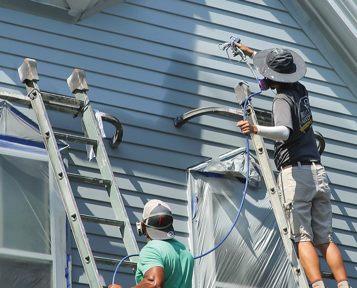 Two workers on ladders use spray paint guns to coat the exterior light blue siding of a building.