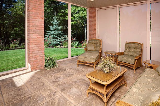 A sunroom with wicker furniture, a coffee table, brick columns, and a view of a lush green backyard.