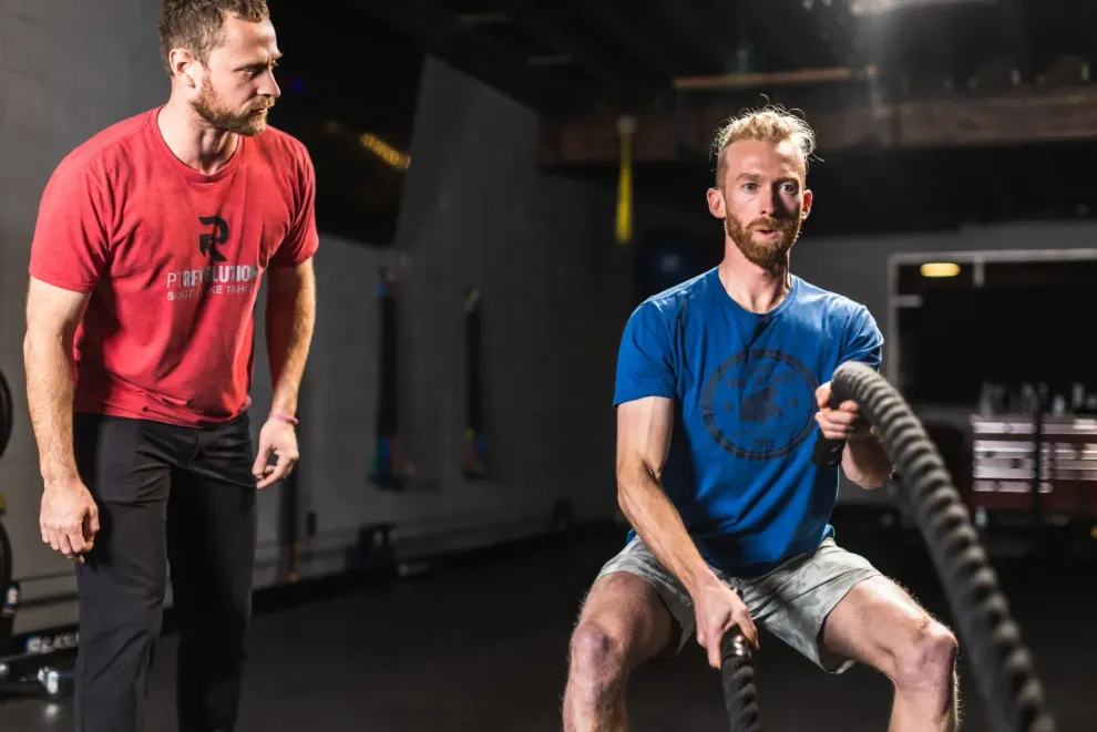 Two men training battle ropes at PT Revolution, one standing in red and one sitting in blue.