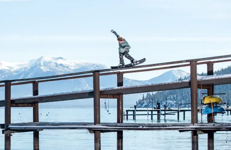 Snowboarder balances on a wooden rail over a frozen lake with snowy mountains in the background.