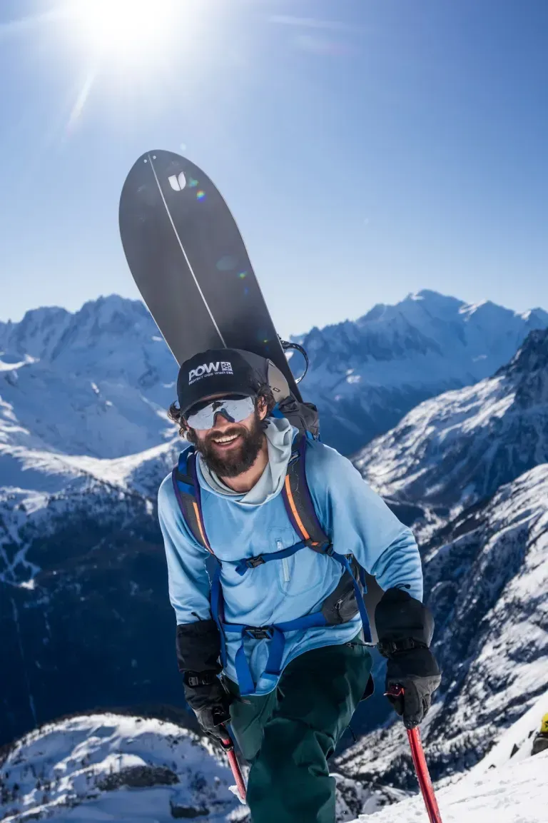 Mountaineer in blue jacket and helmet hiking through snowy alpine mountains with skis on backpack