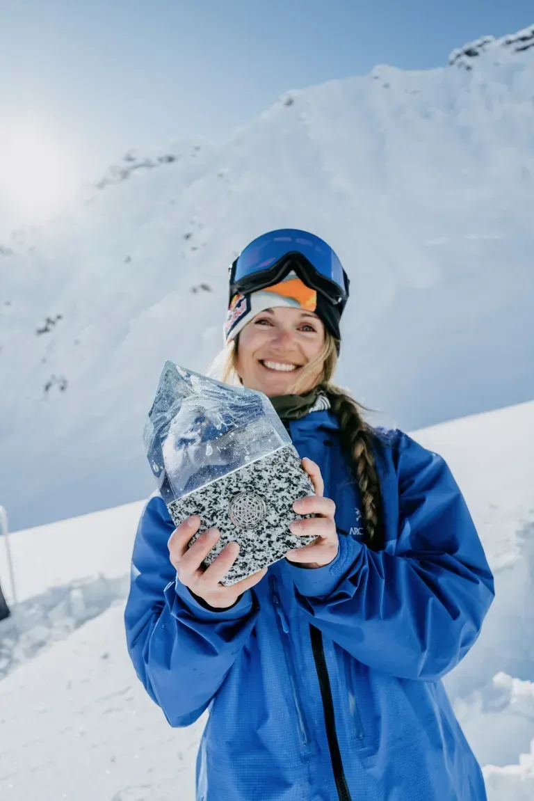 Smiling skier in blue jacket holding a snow-covered rock on a snowy mountain slope