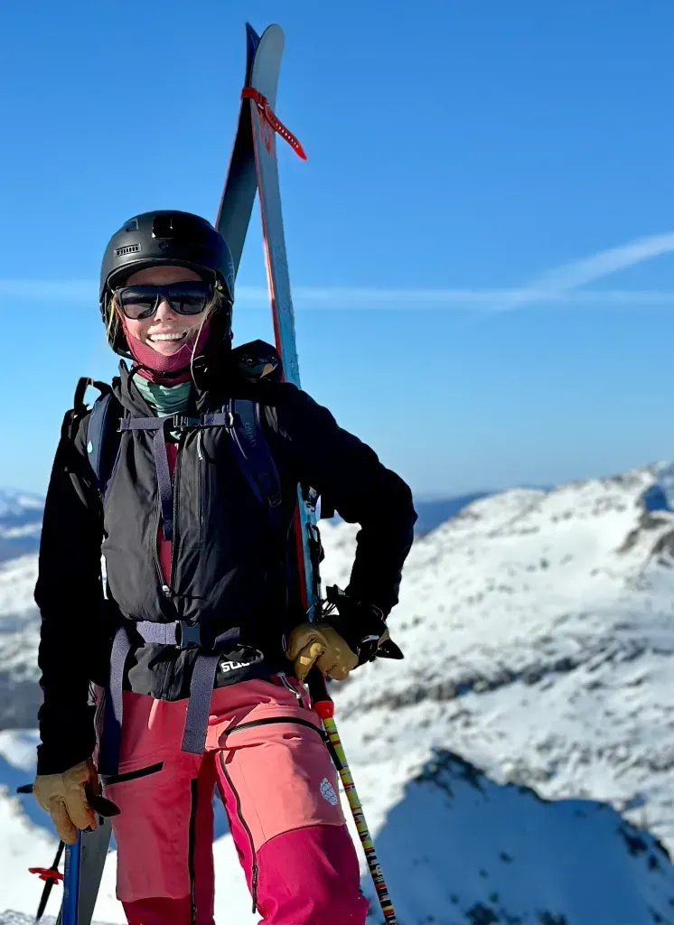 Skier in helmet and goggles standing on a snowy mountain ridge with skis, under a clear blue sky